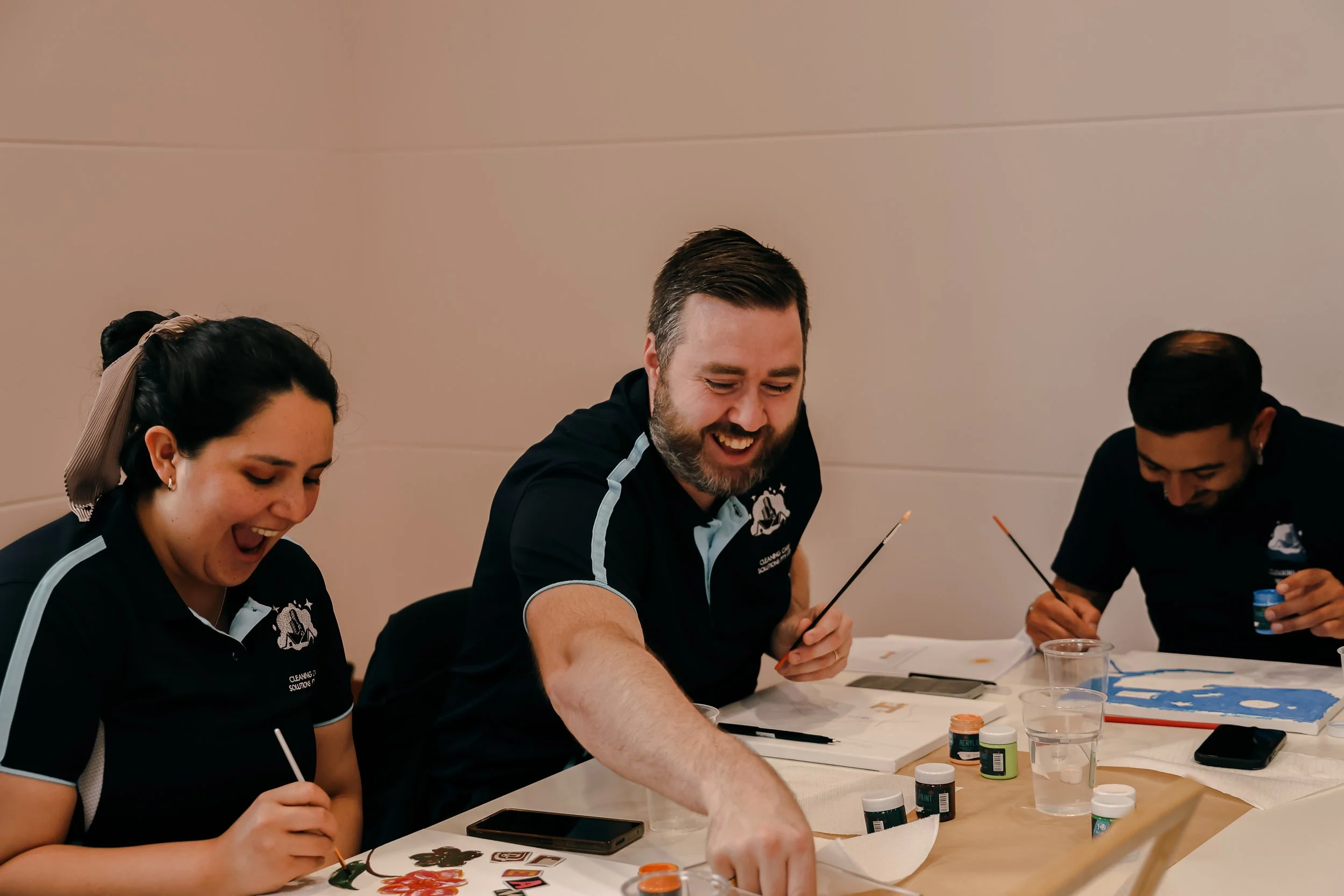 Three people sitting at a table, painting and laughing during an art activity.