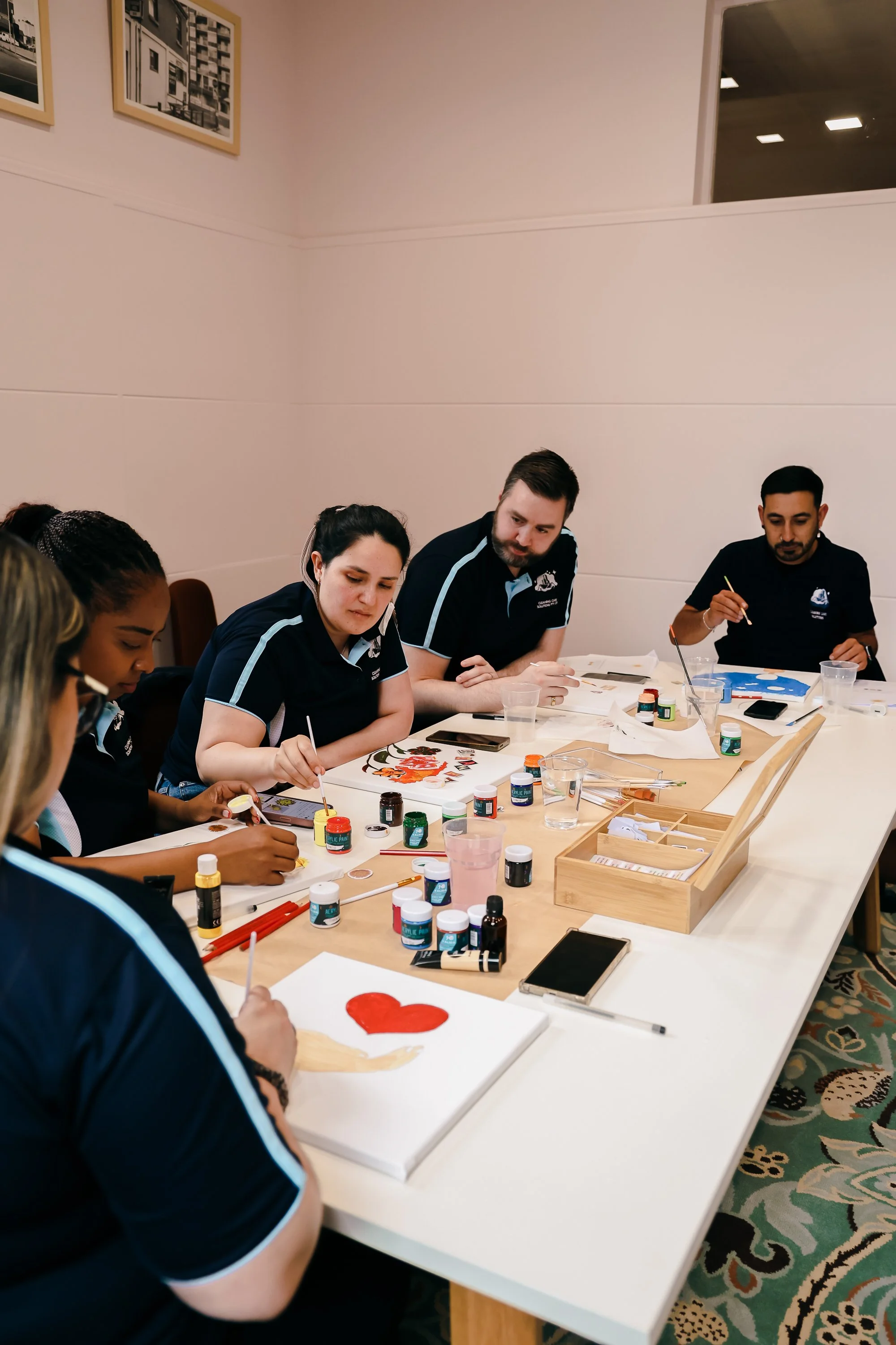 Group of people sitting at a table painting on canvases, with paint supplies and brushes in front of them.