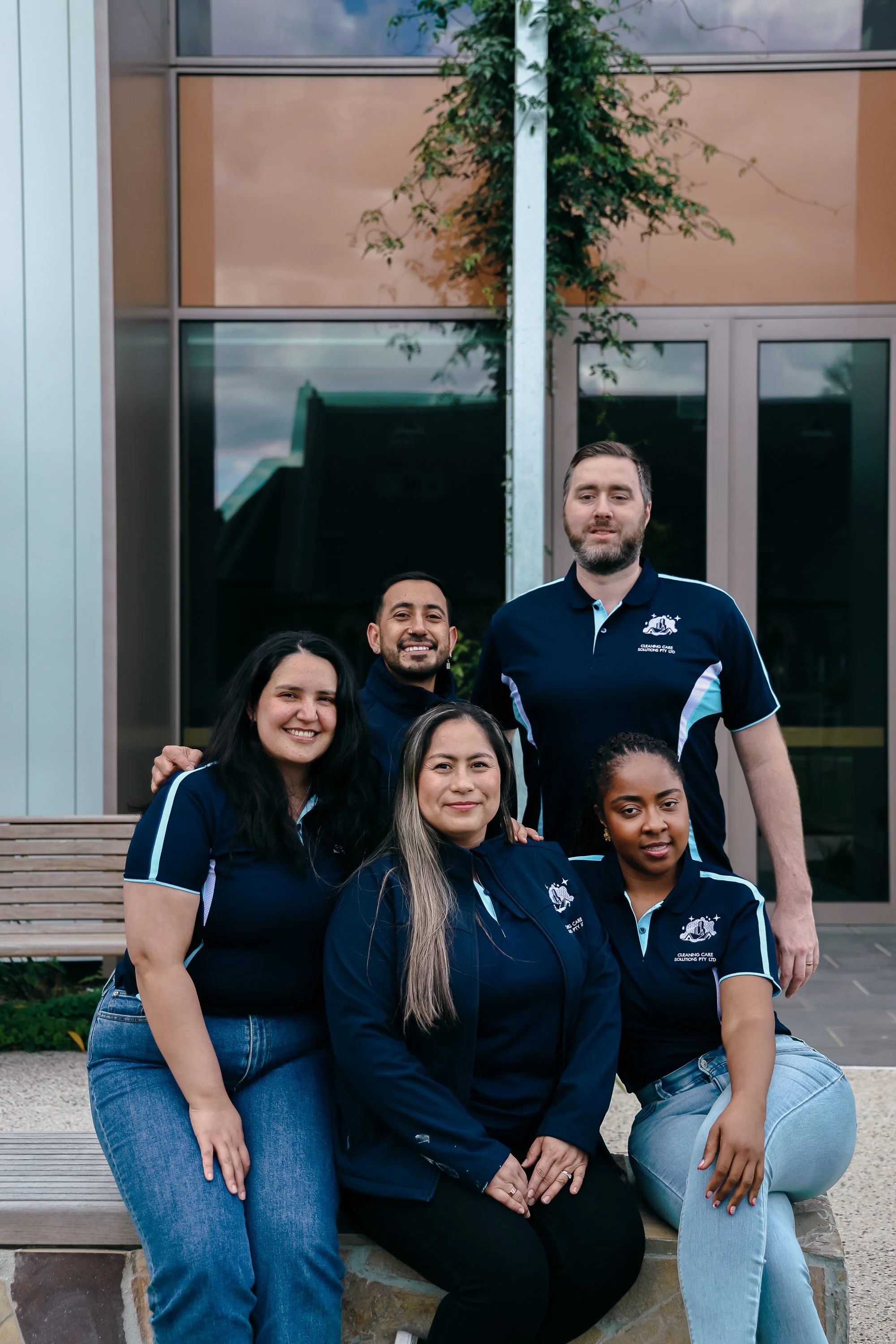 Group of five smiling people in matching navy blue and light blue polo shirts posing outdoors in front of a building with large windows, some seated on a stone bench, others standing behind.