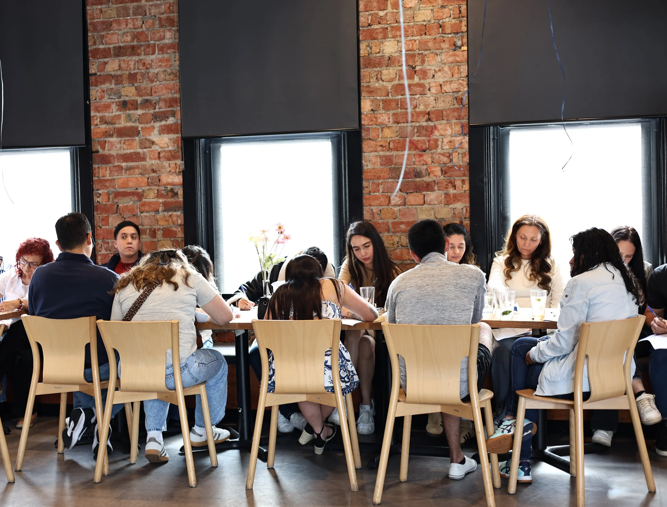 A group of people sitting around a large table in a room with exposed brick walls and large windows, engaged in a meeting or discussion.