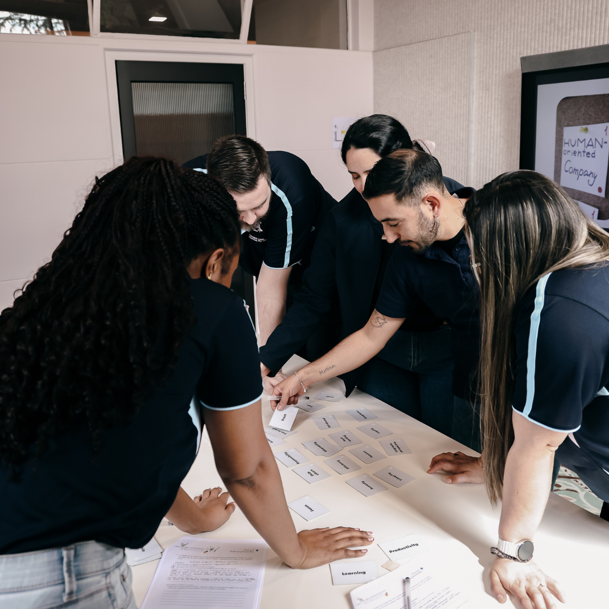 A diverse group of five people collaborating around a table, sorting and discussing paper cards with words on them in an office setting.