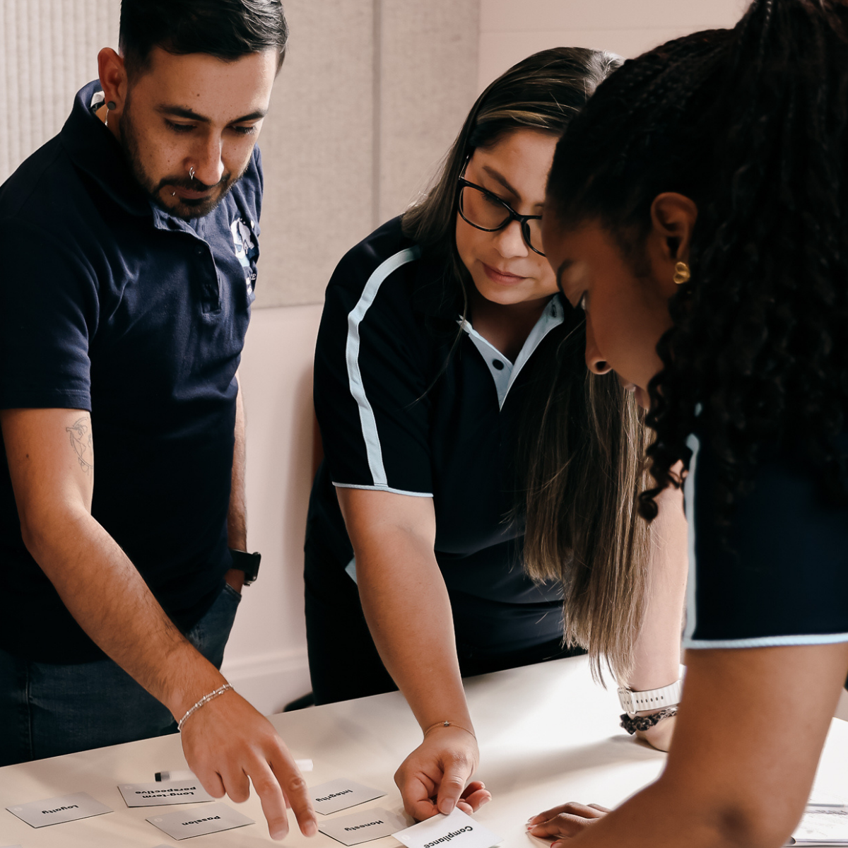 Three people are gathered around a table, looking at and discussing printed cards. They appear to be collaborating or participating in an activity.
