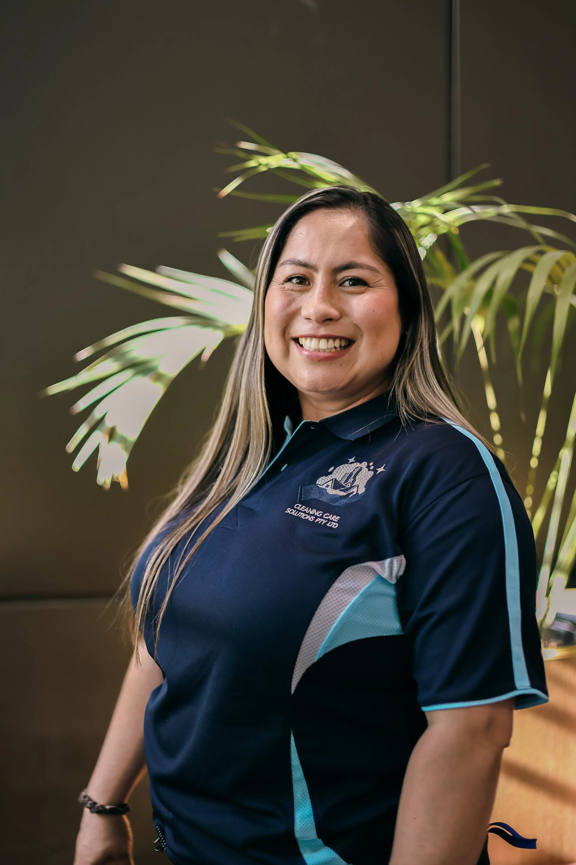 A woman smiling and looking at the camera, wearing a navy blue polo shirt with the logo and text 'Cleaning Care Solutions Pty Ltd.' standing in front of a dark wall and green plant leaves.