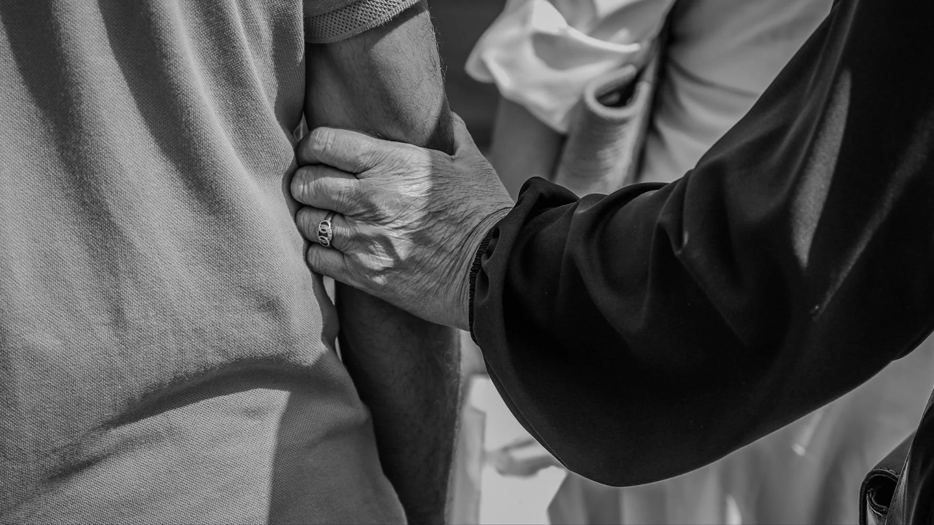 A person wearing a dark jacket is holding the arm of another person wearing a light-colored shirt or jacket. The person holding the arm has an engagement or wedding ring on their finger.