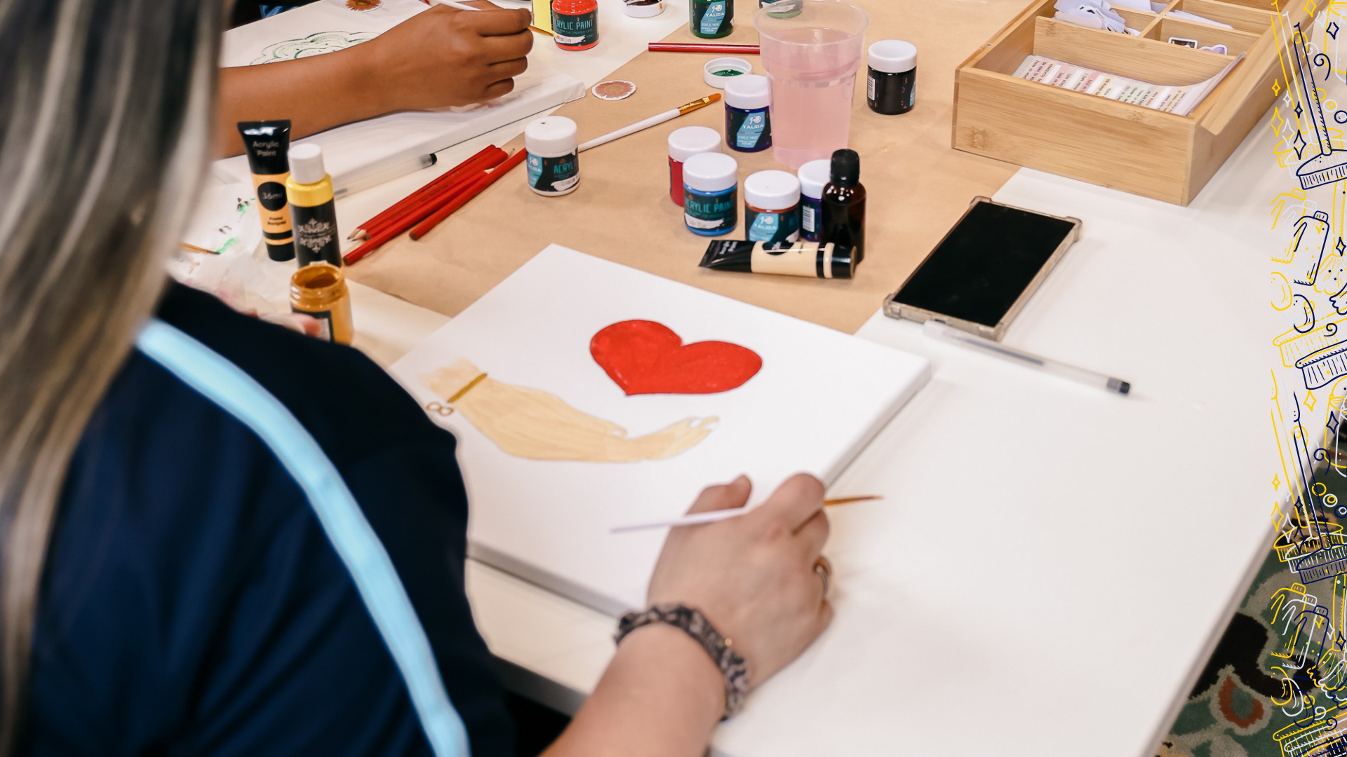 Person painting a picture of a dog with a red heart on a white canvas, surrounded by paint bottles, brushes, and art supplies on a table.