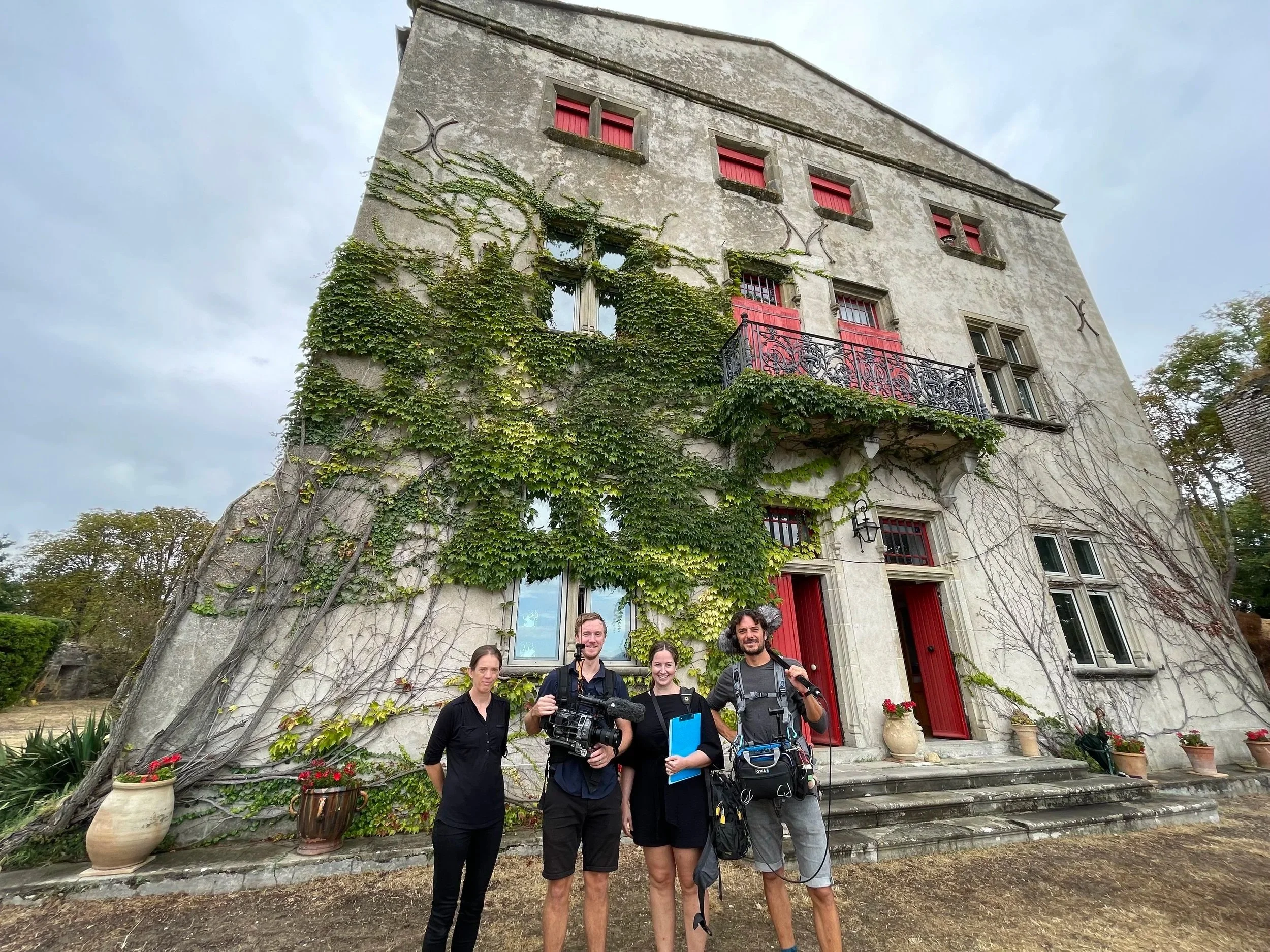 Four people standing in front of a tall, old house with climbing vines, red shutters, and red doors. The house has multiple stories and decorative iron balcony. People are smiling, with one holding a camera and others carrying gear.