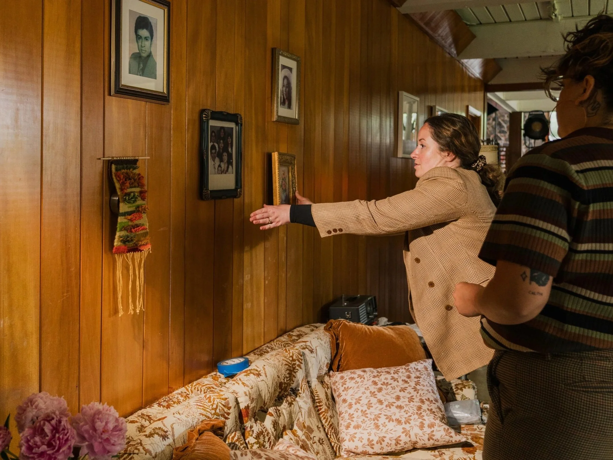 Two women near a wooden-paneled wall with framed photographs and a decorative textile, one reaching toward a framed photo, the other standing beside her.