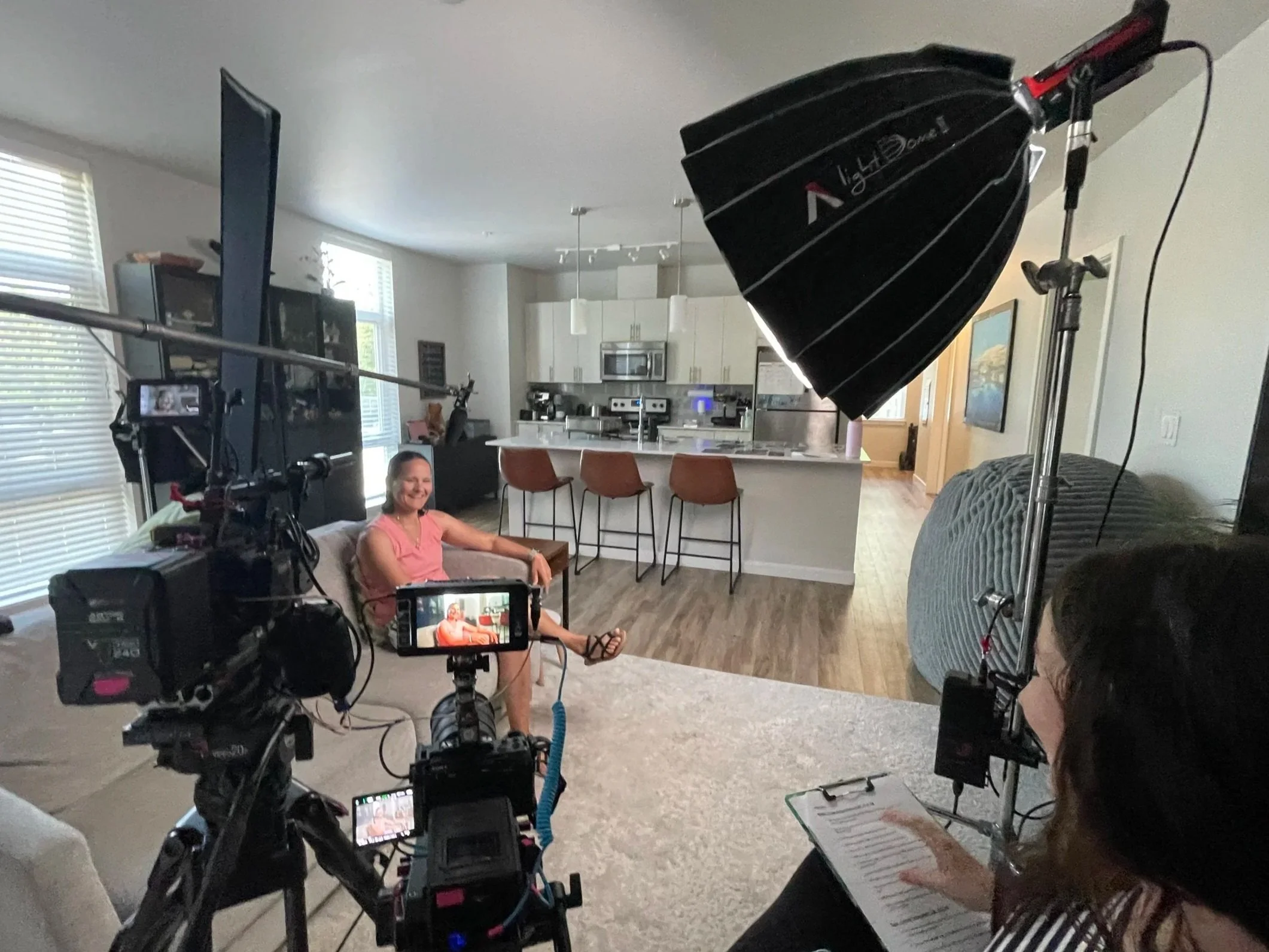 A woman seated on a sofa in a living room, smiling towards a camera, as part of a filmed interview or recording, with lighting equipment and a person with a clipboard nearby. The room has an open kitchen area with bar stools, windows with blinds, and