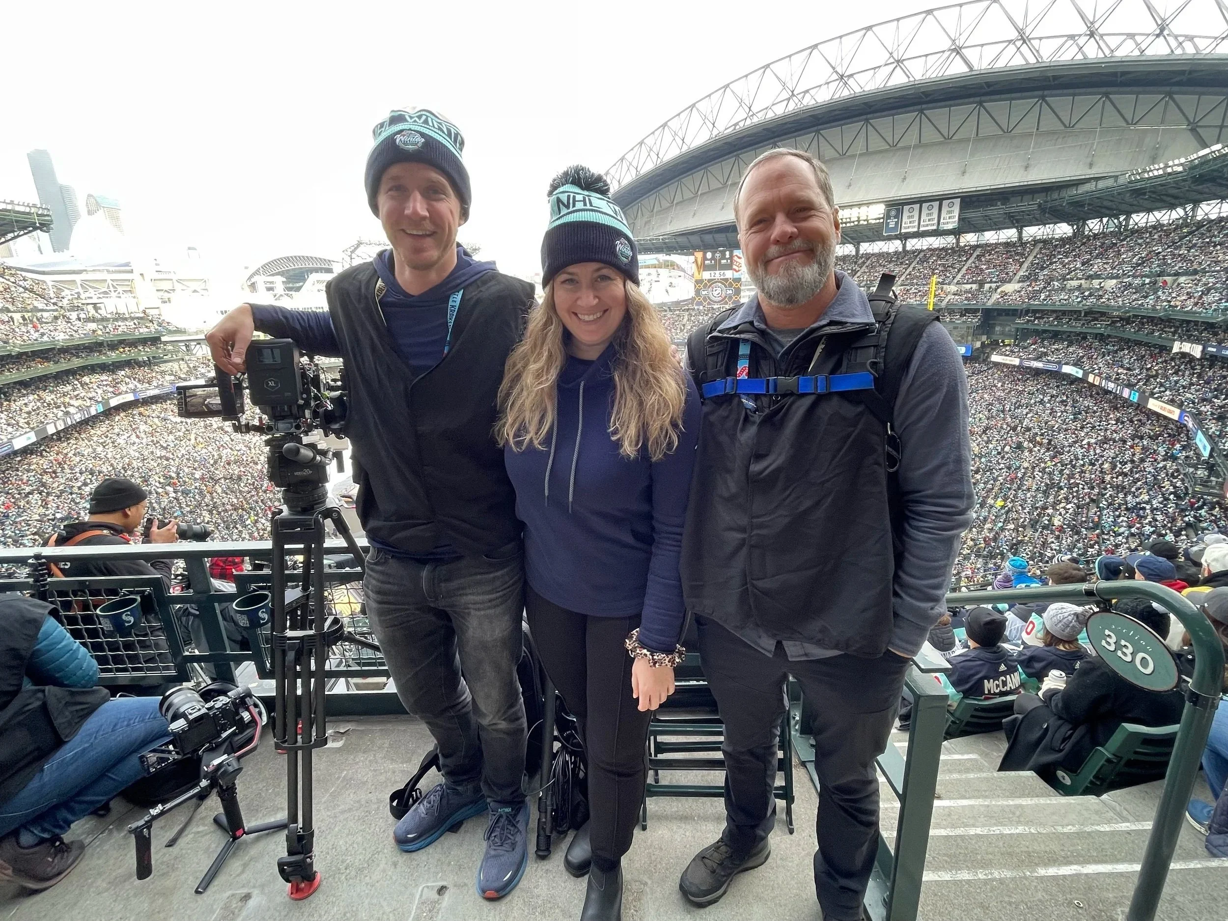 Three people standing together at a stadium, with a large crowd in the background, wearing warm clothing and hats, one person has a camera on a tripod.
