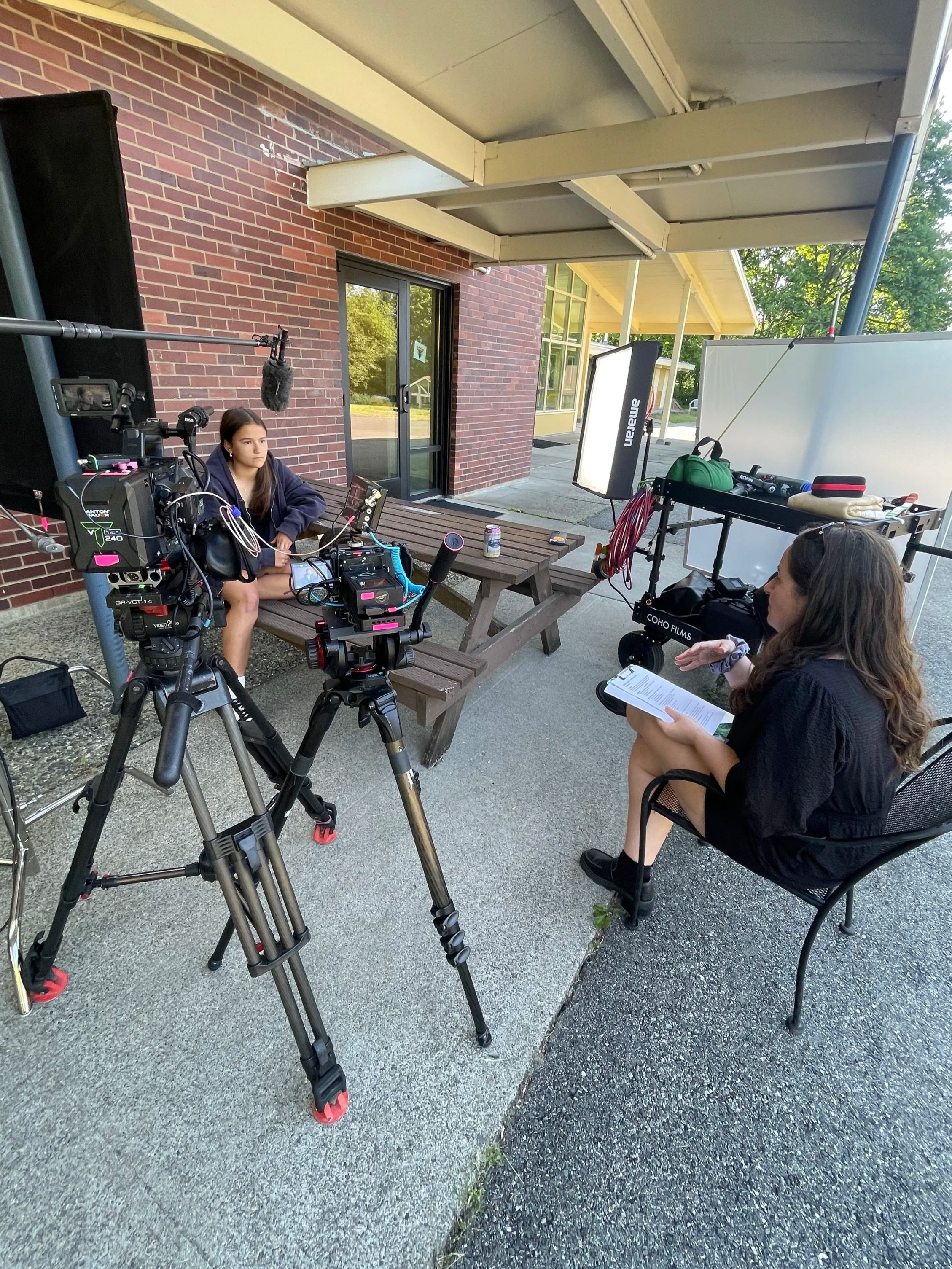 A girl is sitting outdoors, being filmed with professional video equipment, while a woman sits nearby with a script, possibly conducting an interview or recording a video.