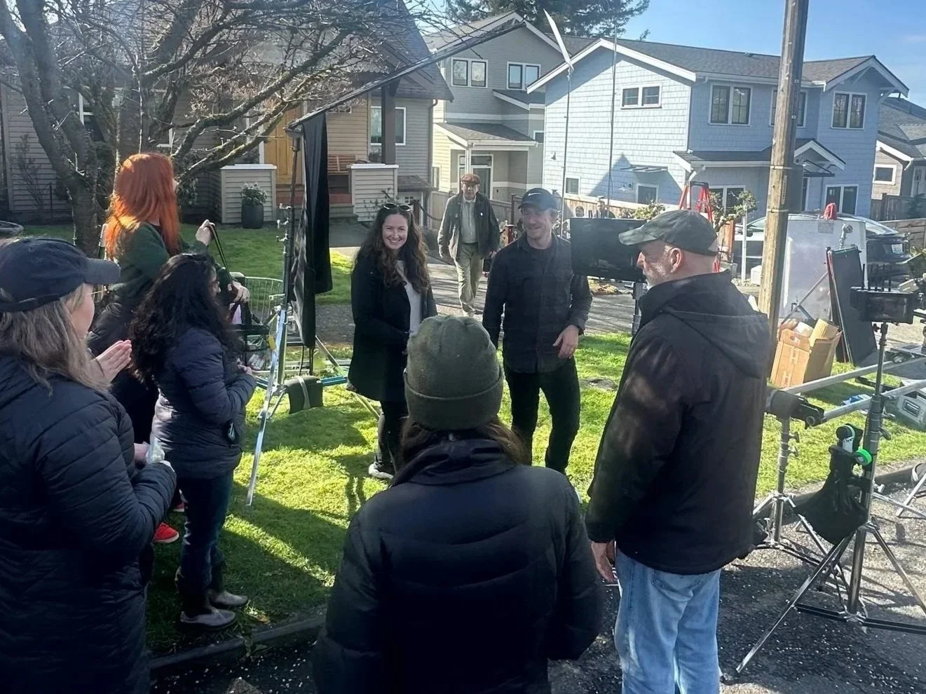 Film crew filming outside on a sunny day with houses and trees in the background, people gathered around cameras and equipment.