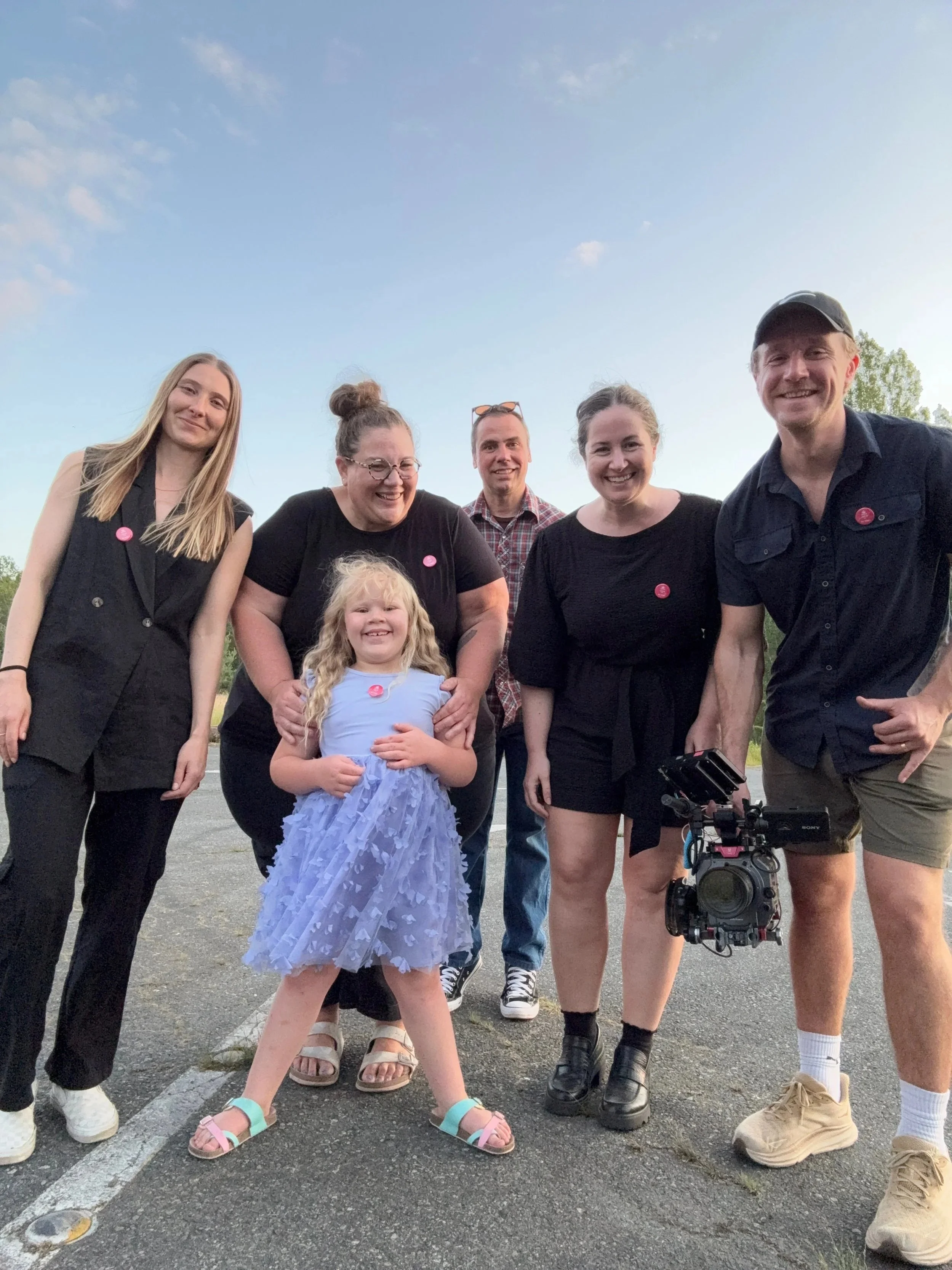 Group of six people, including a young girl in a light purple dress, standing outdoors in a parking lot during the evening under a partly cloudy sky.