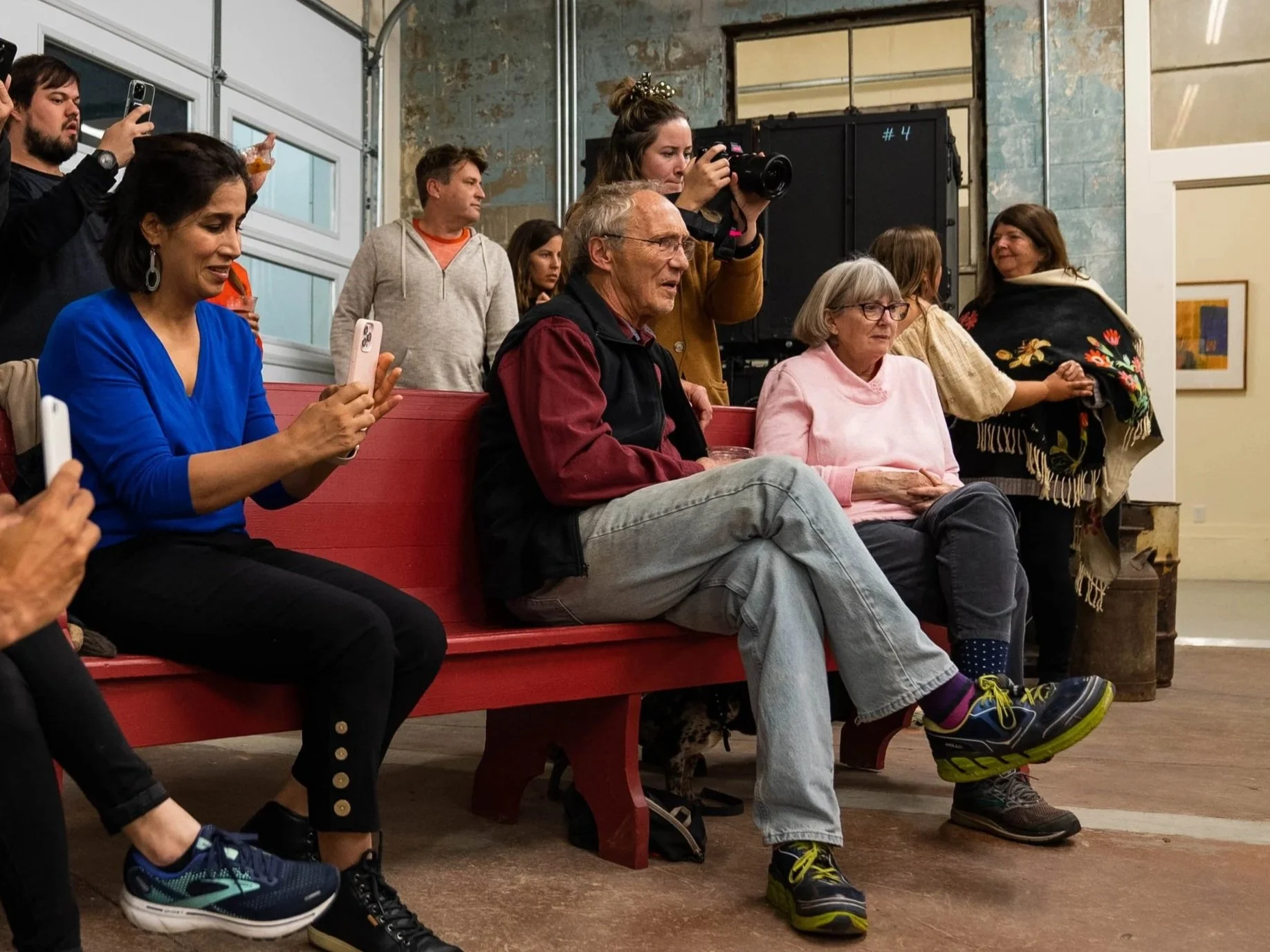 Group of people sitting and standing around a red bench in an art gallery or indoor event space, with one woman taking a photo and others watching or waiting, some holding phones, various ages and styles, exposed walls, and framed artwork in the back