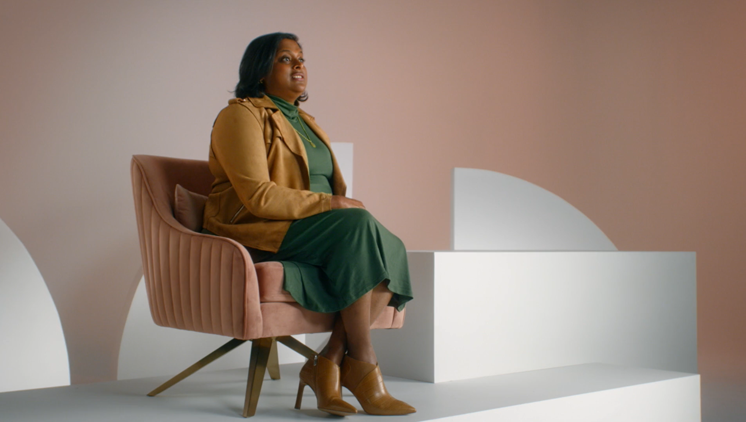A woman sitting in a pink velvet armchair with a brown leather jacket, green dress, beige ankle boots, and a necklace, against a minimalist background with geometric shapes.