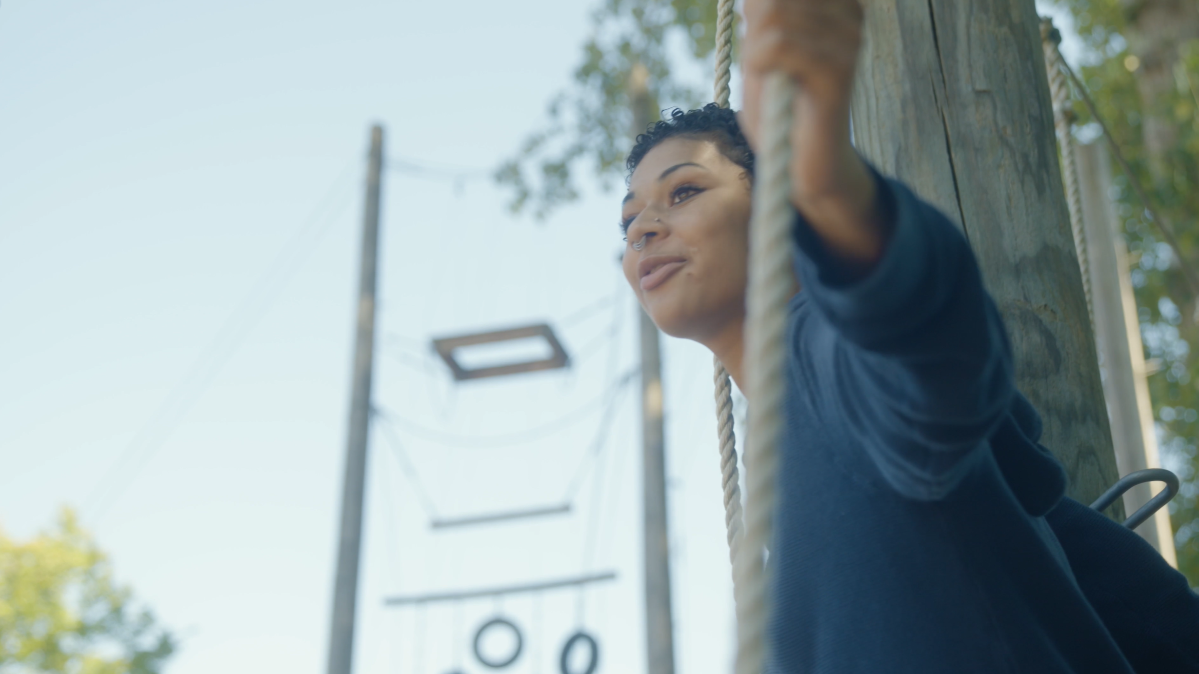 A woman with short curly hair and nose piercings leans against a wooden pole at an outdoor ropes course, looking thoughtfully into the distance with a clear blue sky in the background.