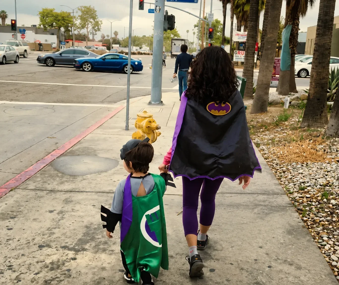 Mom dressed as Batgirl and son as superhero walk hand in hand along a city sidewalk.