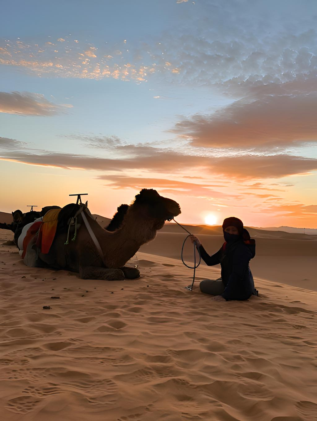Rosalie sitting beside a camel in the Sahara Desert at sunset, reflecting curiosity, exploration, and cultural connection.