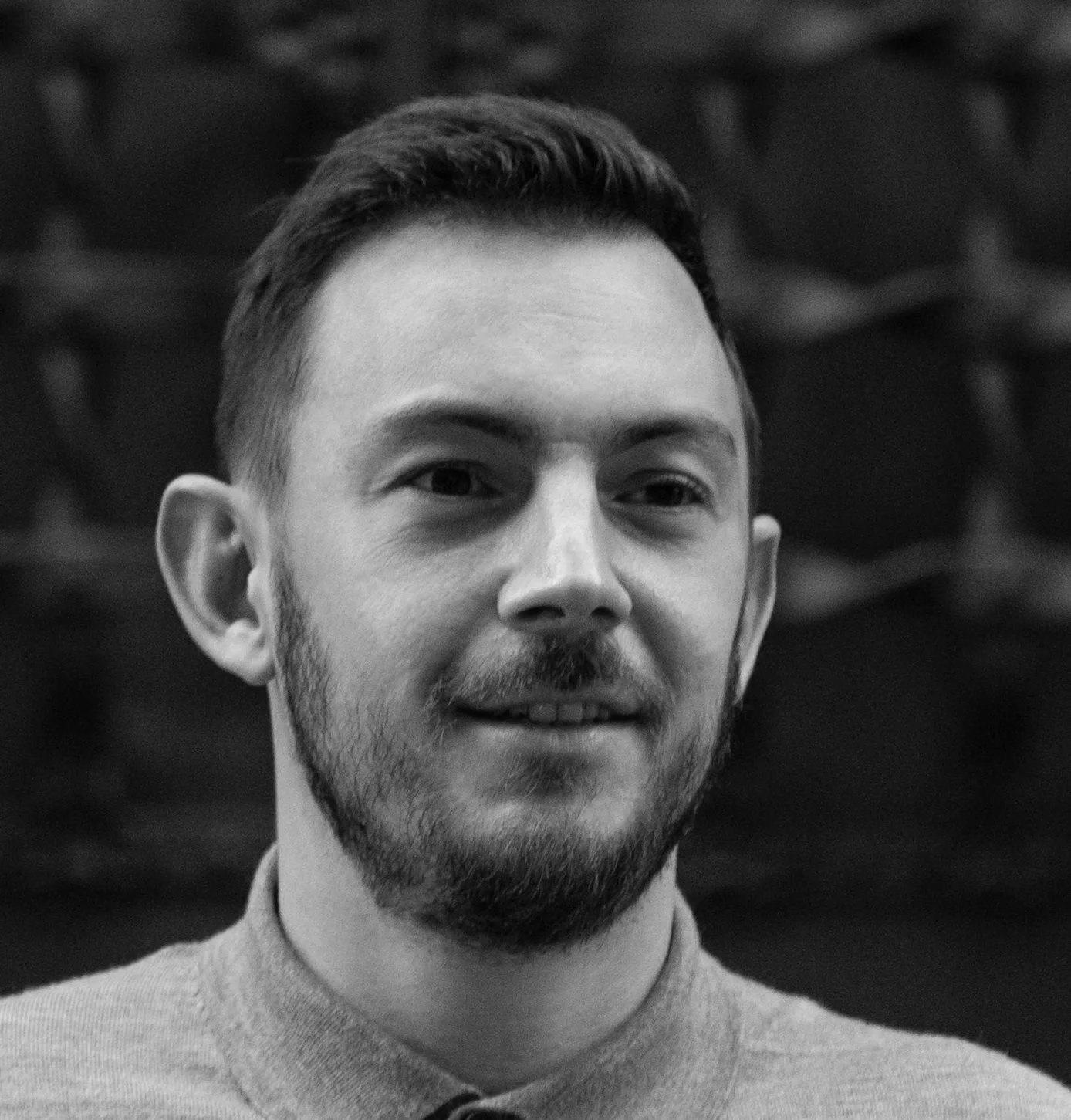 Black and white portrait of Derek Main with short hair, beard, and mustache, wearing a collared shirt, looking slightly to the side.