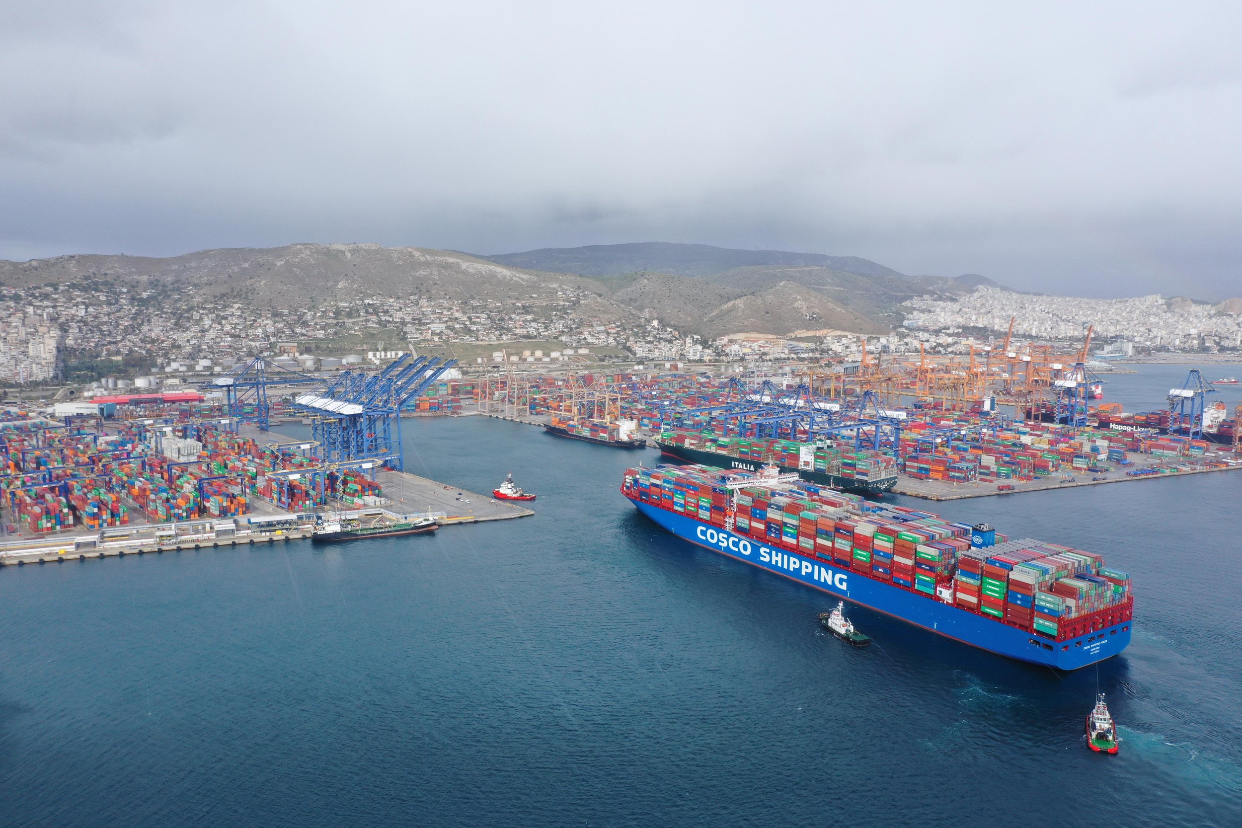 Large cargo ship labeled 'COSCO SHIPPING' sailing through a busy port filled with stacked containers and multiple cranes, with hills and a city in the background.