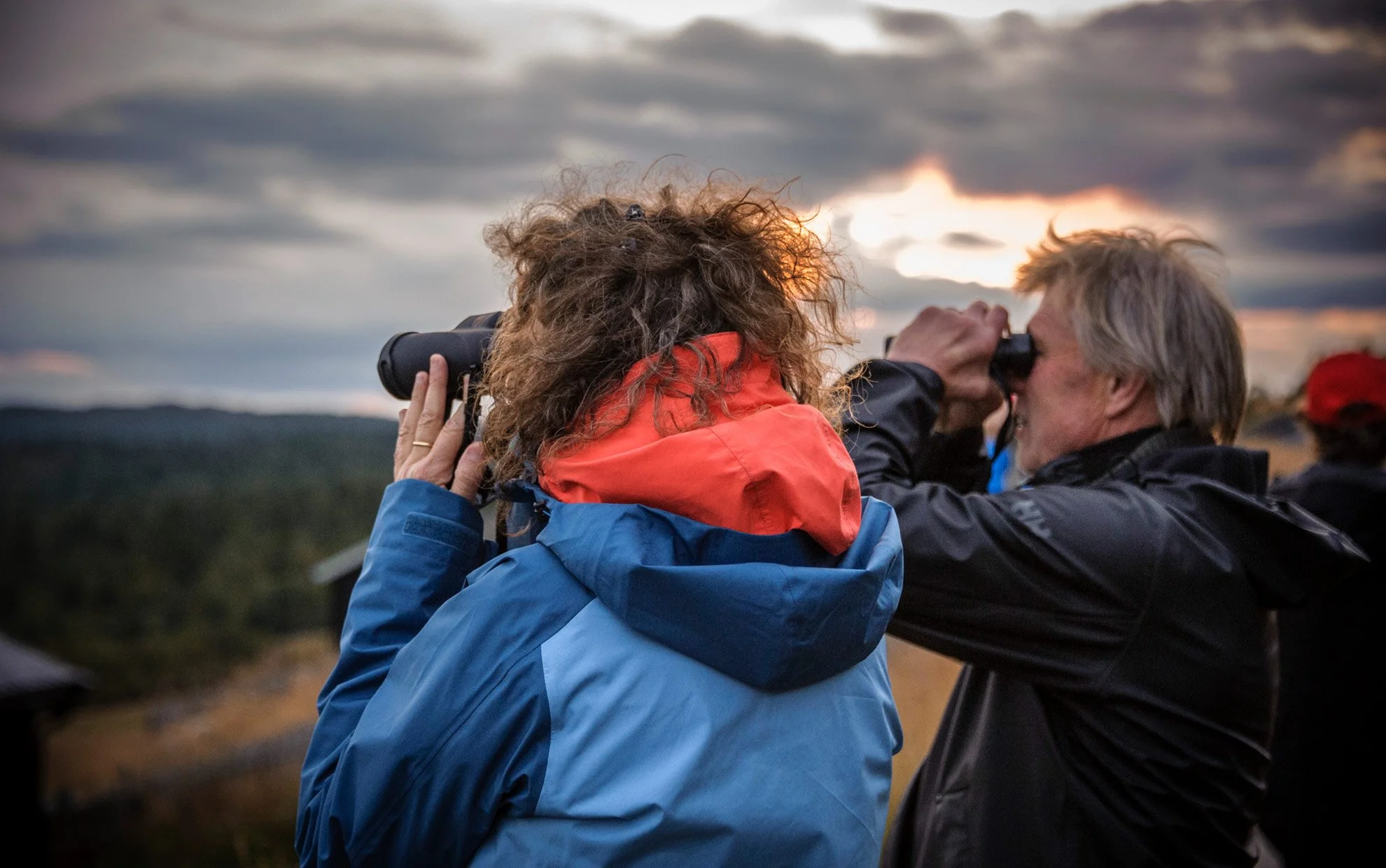 Two people outdoors, one pointing through binoculars while the other looks at the horizon during sunset.