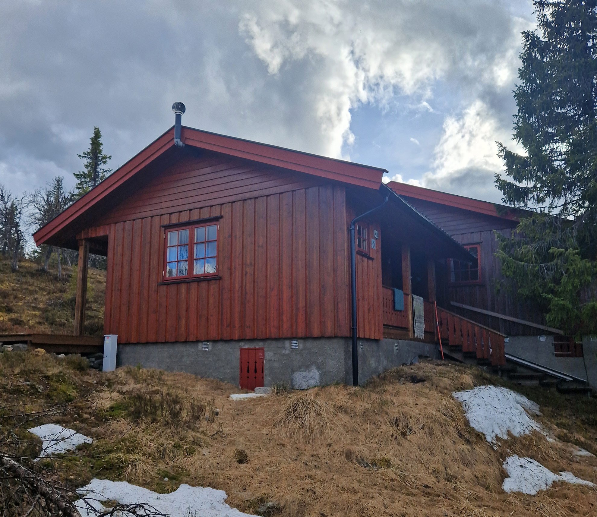A wooden cabin with a red roof on a hillside, surrounded by trees and under cloudy sky.