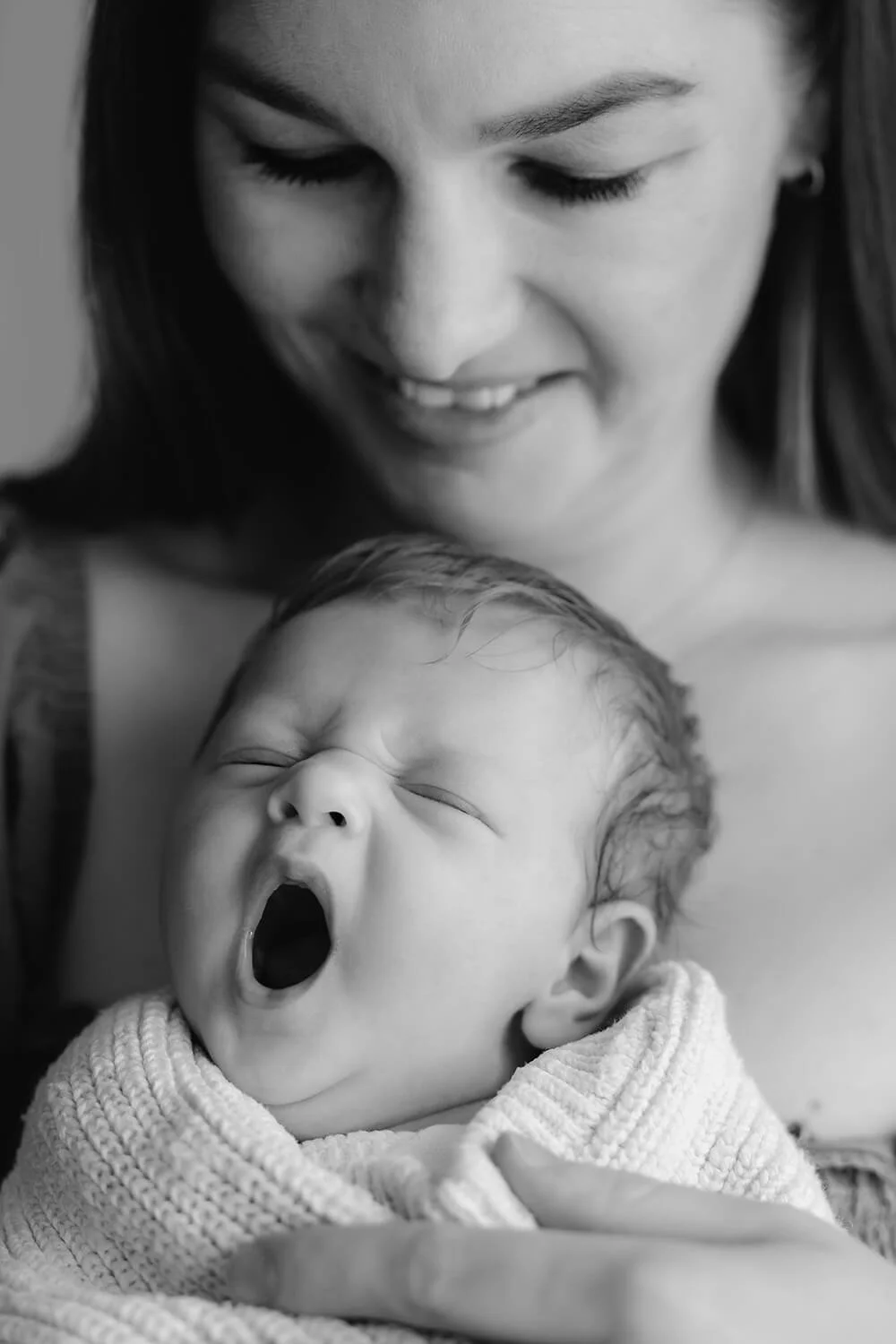 A woman with long dark hair looks down smiling at a yawning newborn wrapped in a blanket.