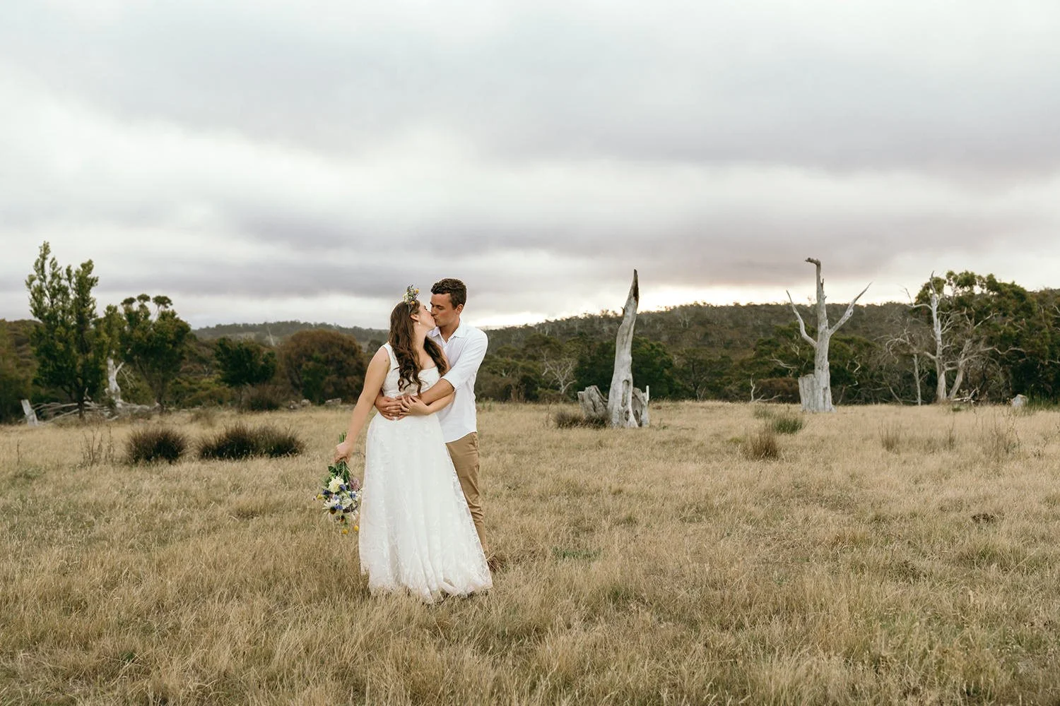 A couple in wedding attire standing in a grassy field, kissing. The woman holds a bouquet of flowers, and the man is wearing a white shirt and tan pants. The background features sparse trees and a cloudy sky.