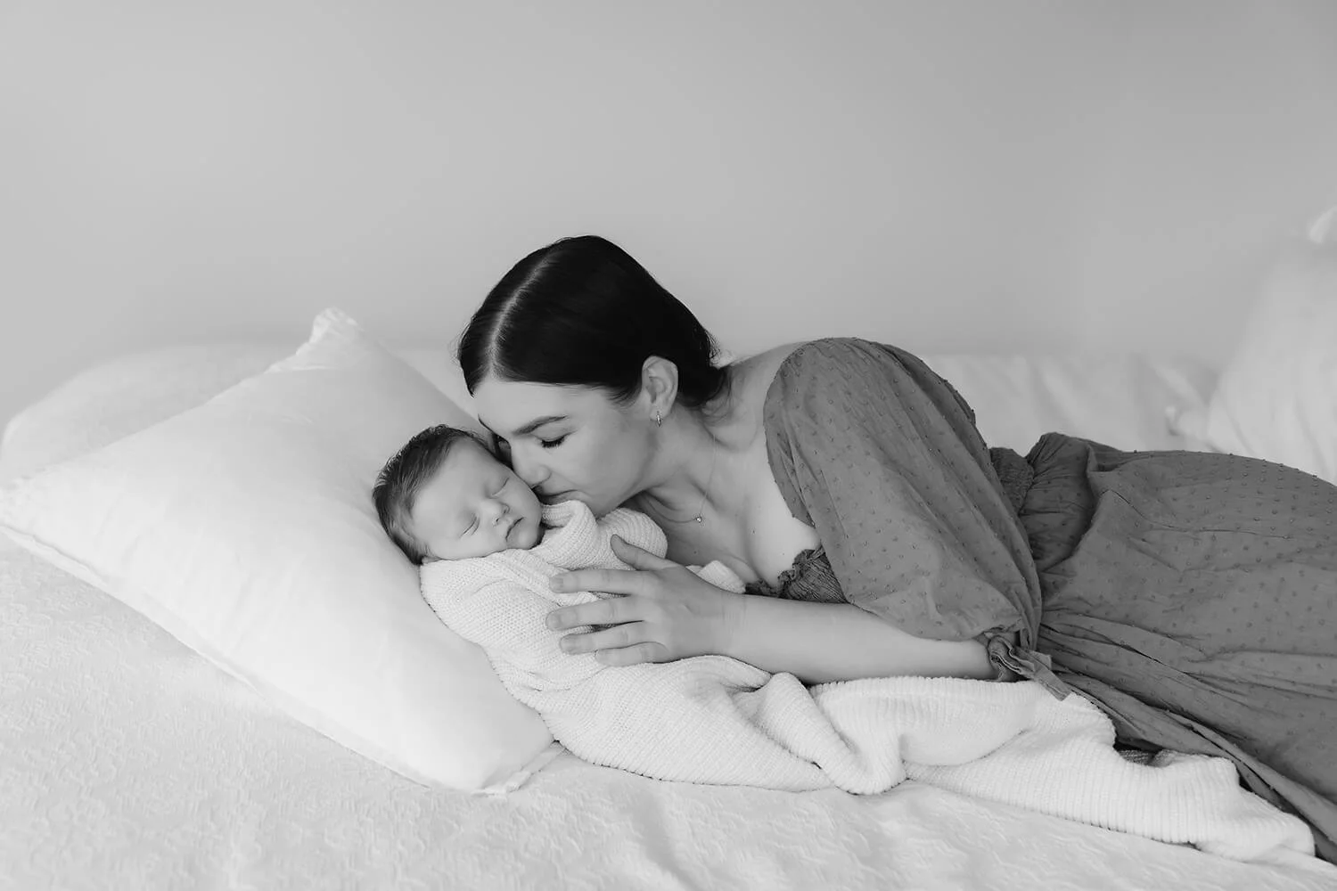 A woman lying on a bed, gently kissing a sleeping baby's forehead.