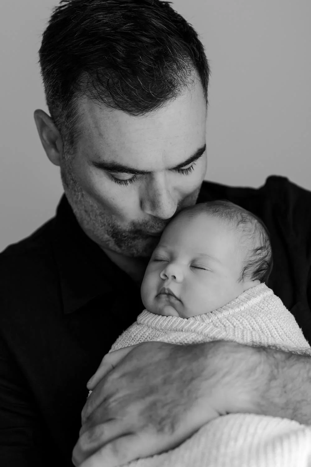 A black and white photo of a man holding a sleeping baby close to his chest, with the man gently touching the baby's head.