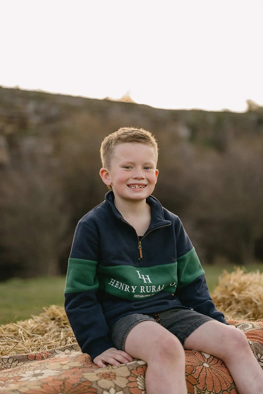 A smiling young boy with short blonde hair sitting outdoors on a blanket during daytime in a rural setting.