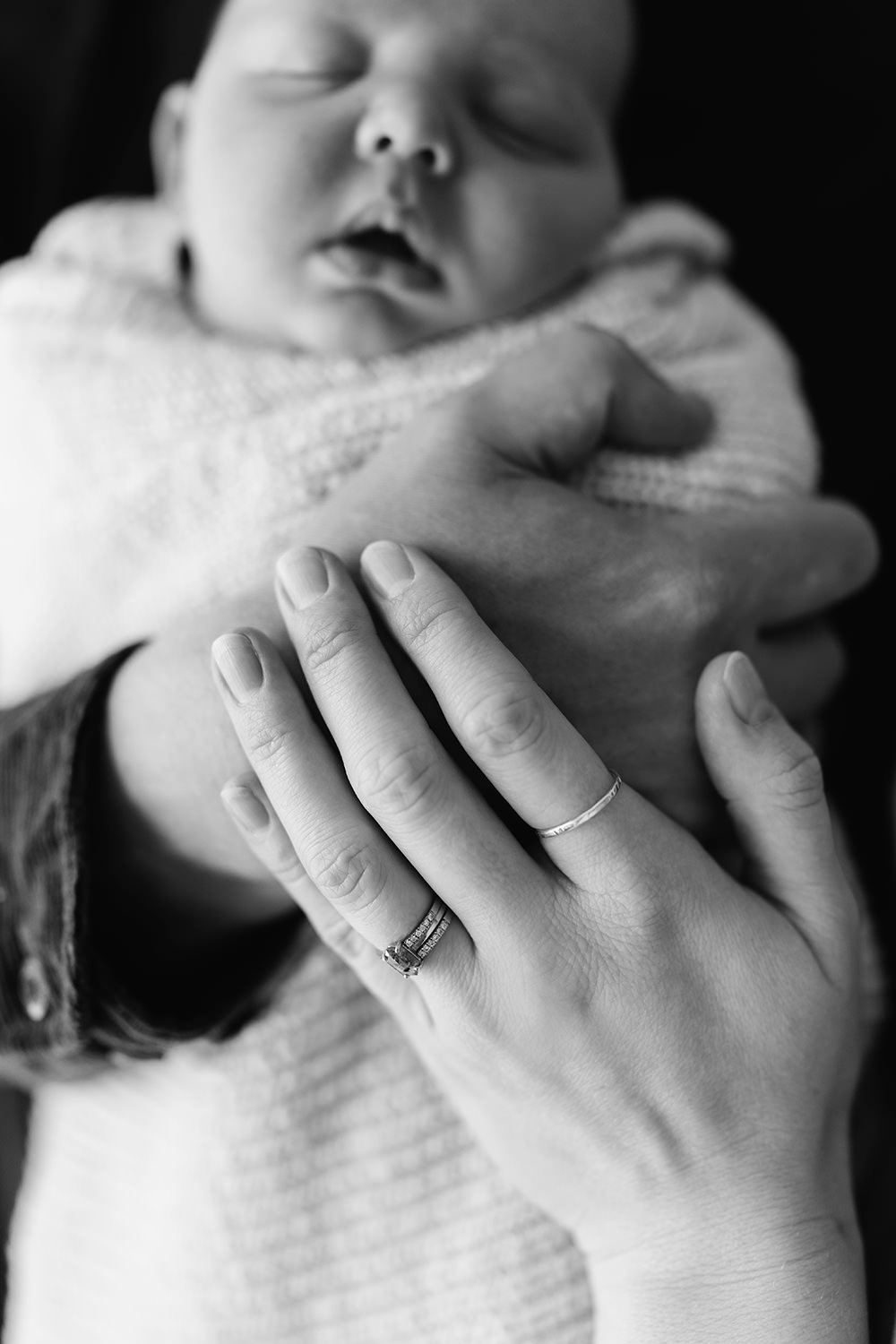 Close-up of a newborn baby being held by an adult, black-and-white photograph, focusing on the baby's face and the adult's hand with rings.
