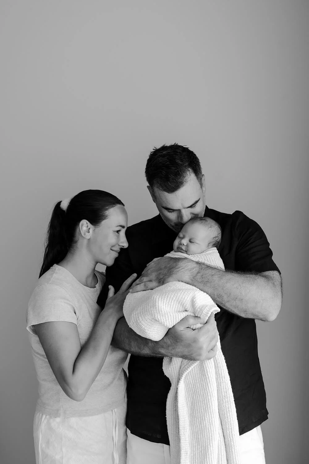 Black and white photo of a family holding a newborn baby. The mother and father are close to each other, with the mother touching the baby's head and the father looking down at the infant.