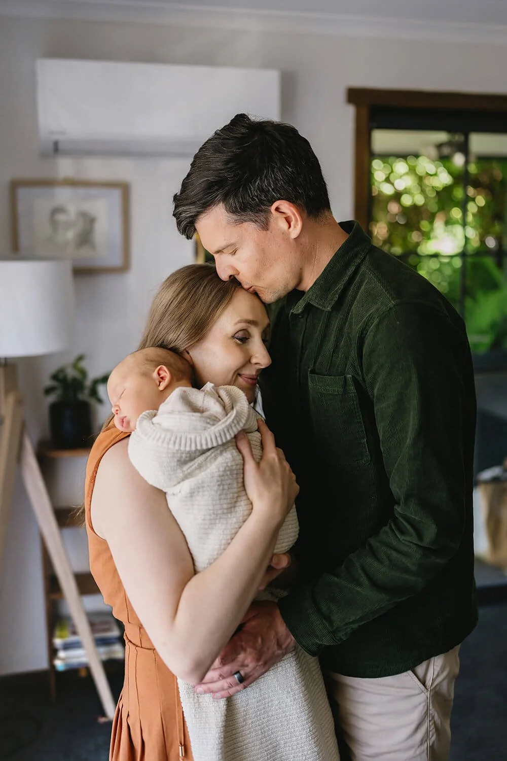 A family with a mother, father, and newborn baby sharing a tender moment indoors. The mother is holding the sleeping baby close to her chest, while the father gently rests his forehead on the mother's head, creating an intimate and loving scene.