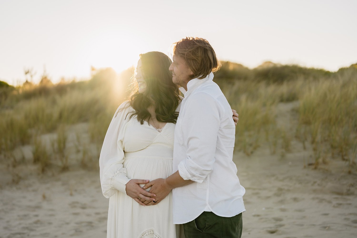 A happy couple standing on the beach during sunset, embracing and smiling at each other.