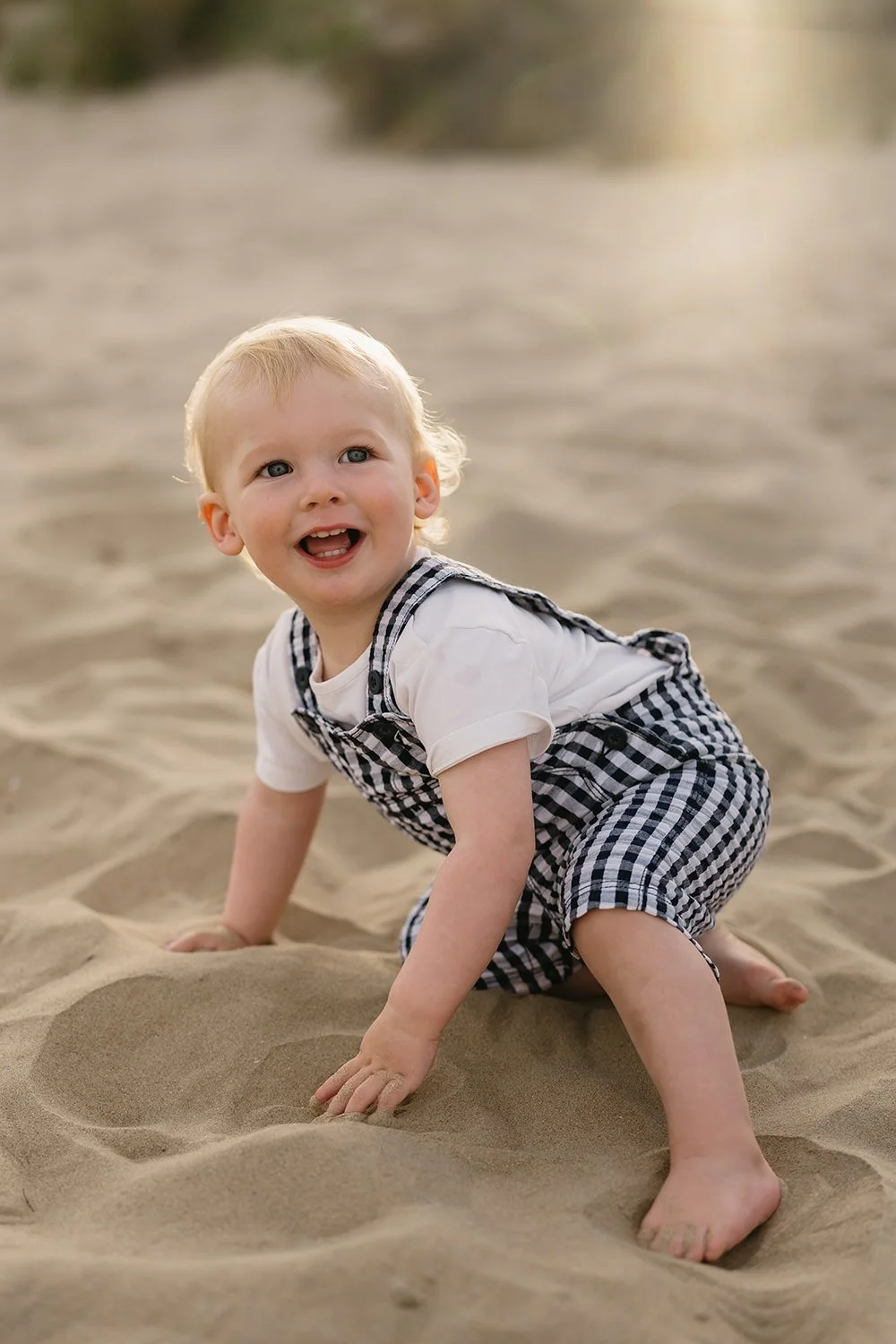 A young child with blonde hair and blue eyes, wearing a white shirt and checkered overalls, crawling on sandy ground at a beach or desert, smiling and looking upward.