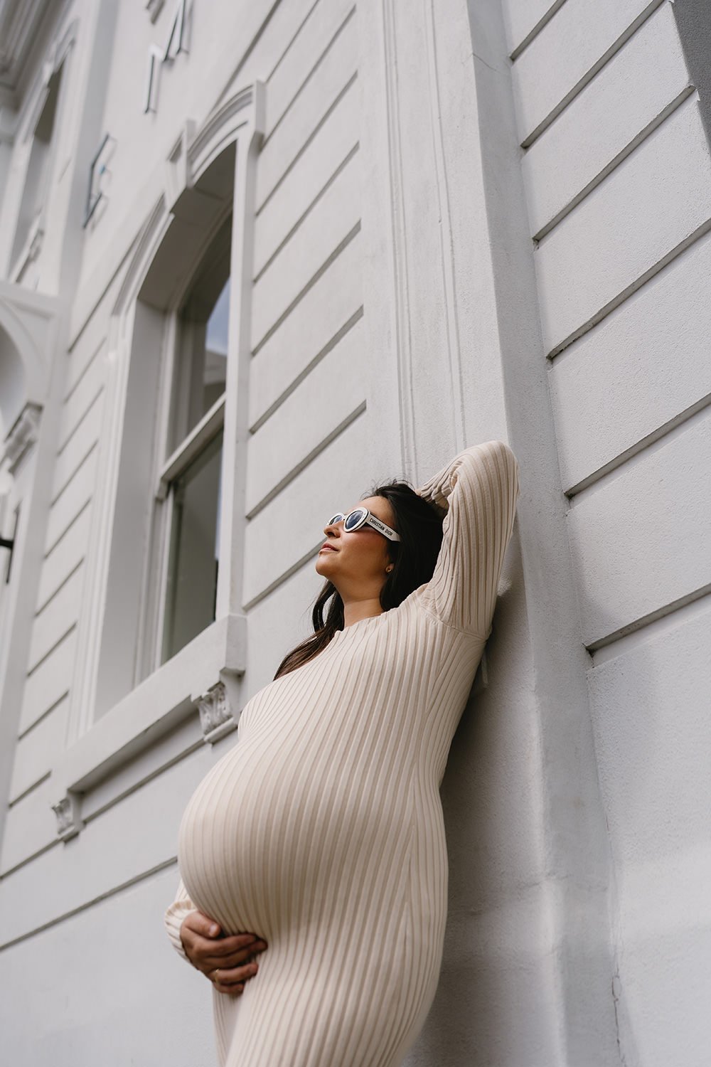 A pregnant woman leaning against a wall outside, wearing sunglasses and a beige ribbed dress, with one hand on her belly and the other behind her head.