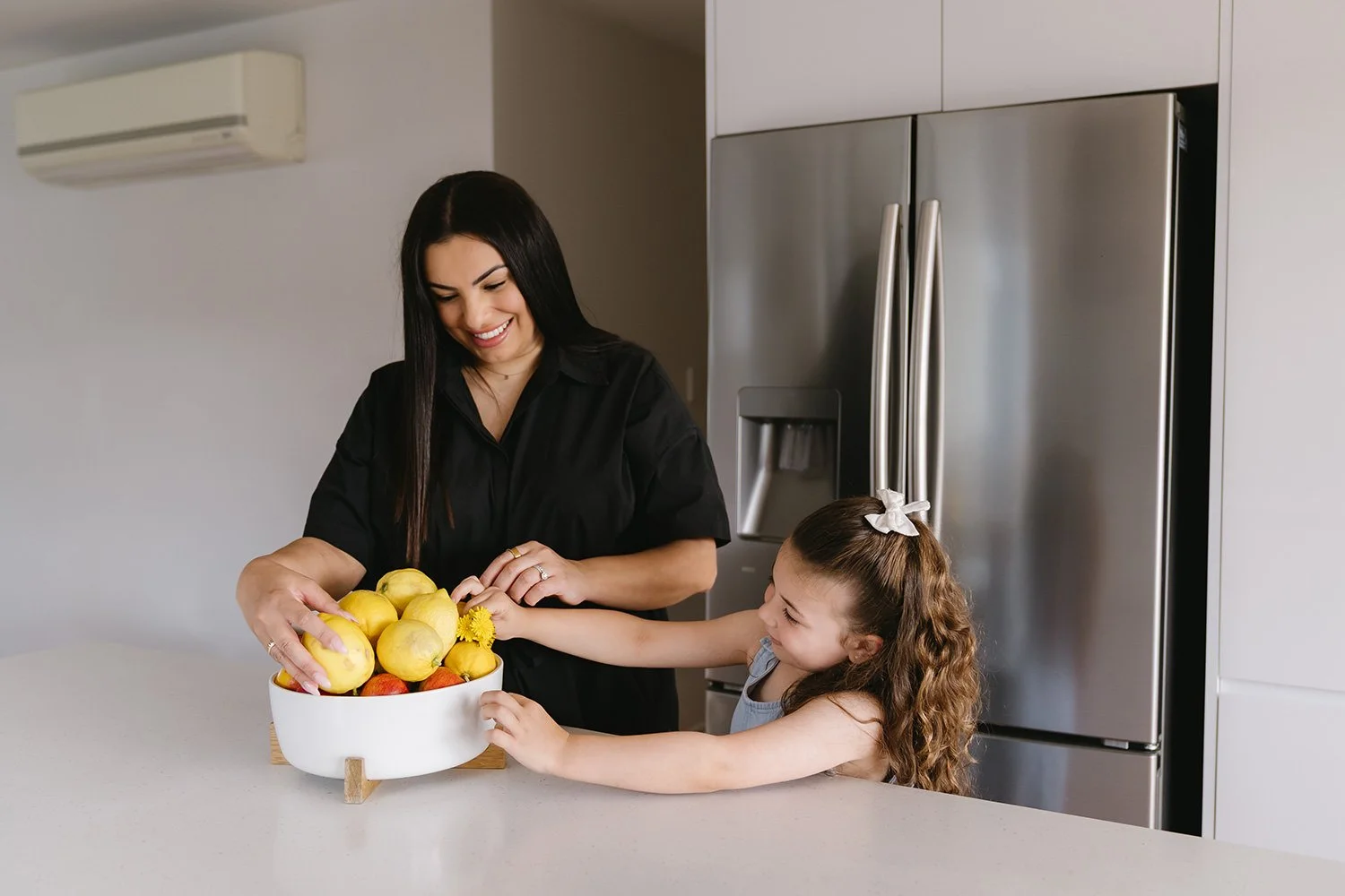 A woman and a girl sharing a laugh while reaching for a bowl of apples and lemons on a kitchen counter.