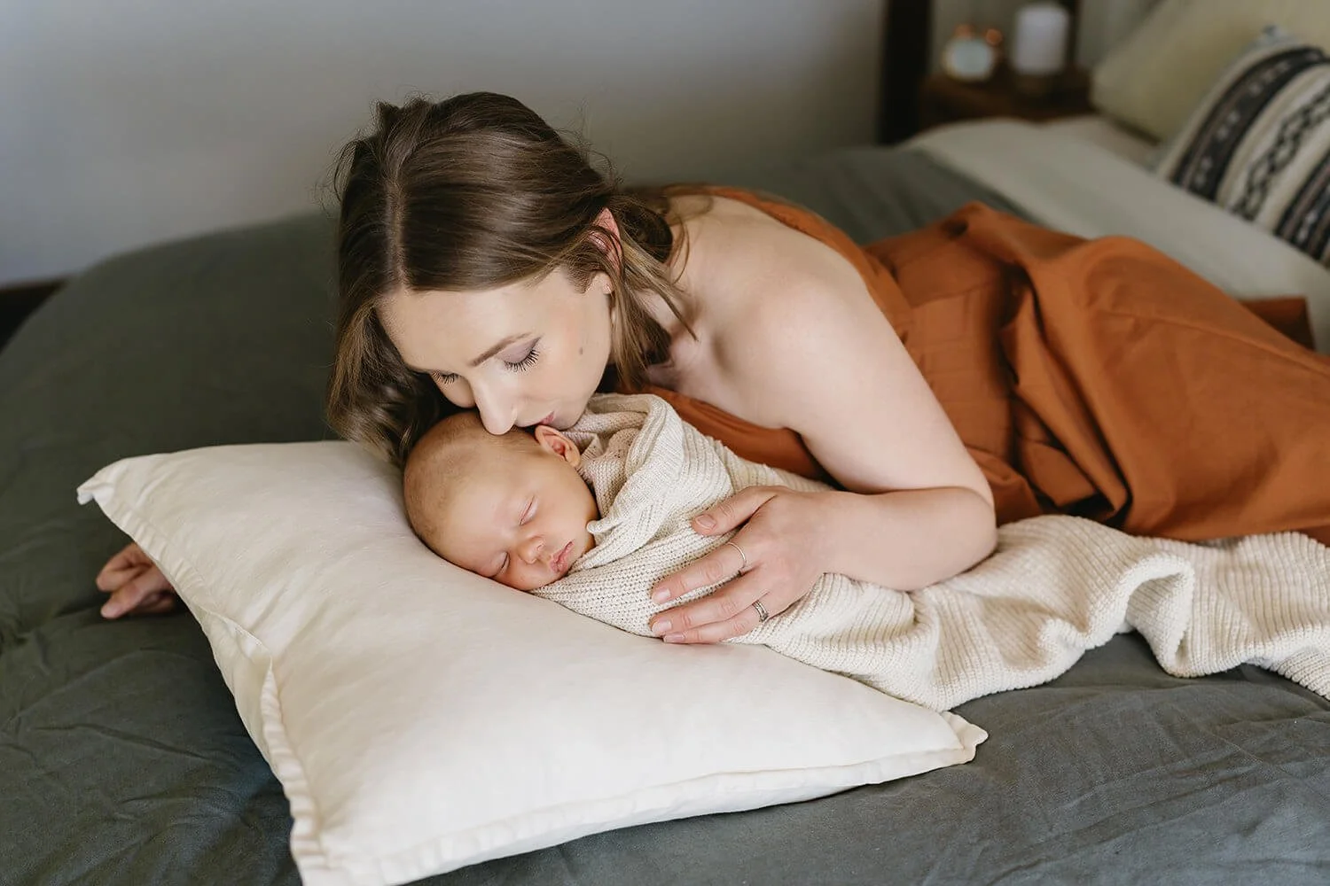 A woman with brown hair is lying on a bed, tenderly kissing a sleeping baby who is wrapped in a beige blanket.