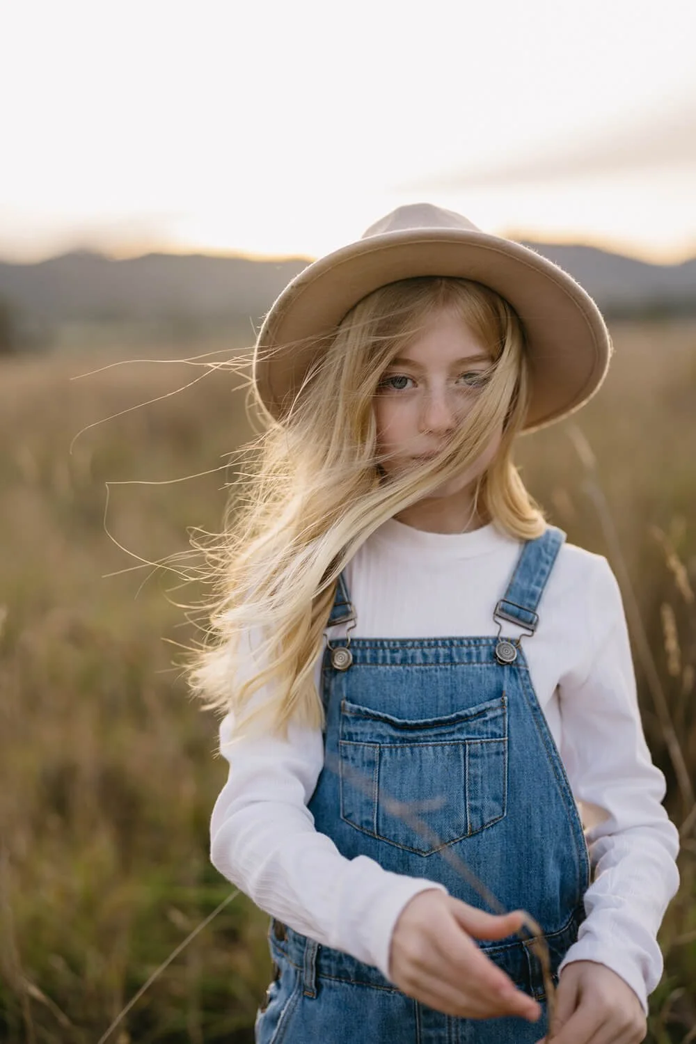 A young girl with long blonde hair, wearing a beige hat, white shirt, and denim overalls, standing in a field during sunset.