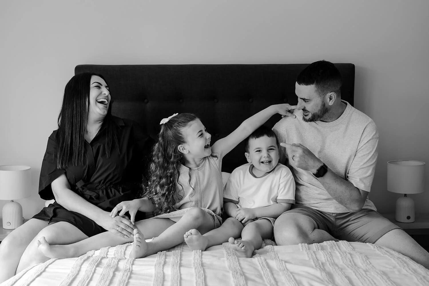 Family of four, two children and two adults, sitting on a bed, laughing and playing together in a black and white photo.