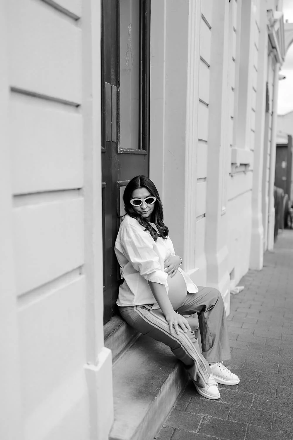 A pregnant woman with long hair wearing white sunglasses, a white loose shirt, striped pants, and white sneakers, sitting on a step outside a building, holding her pregnant belly.