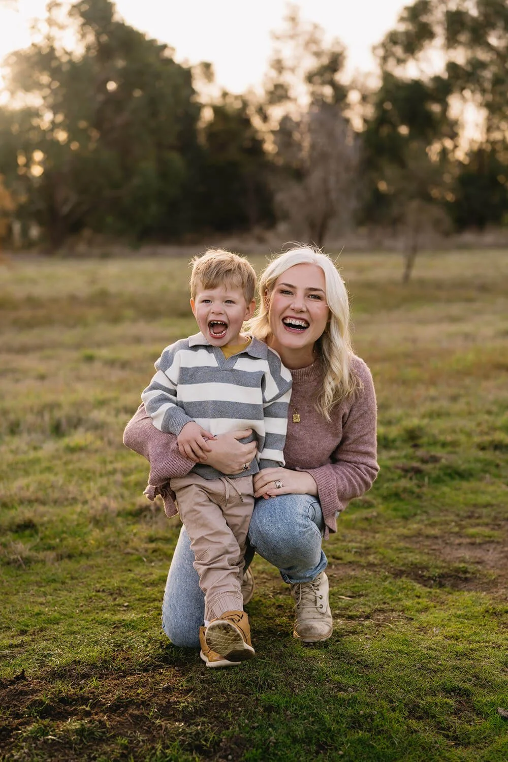 A woman kneeling outdoors in a grassy field, holding a young boy, both smiling and laughing, during sunset with trees in the background.