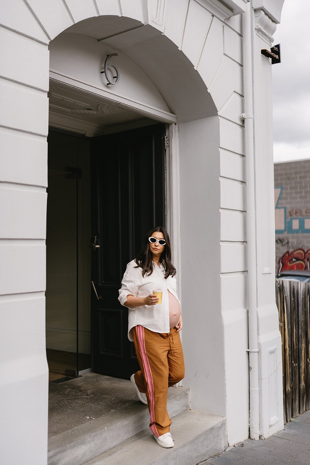 Pregnant woman with white sunglasses, white shirt, tan pants with pink stripes, and white sneakers standing on a step outside a white building, holding a yellow cup.