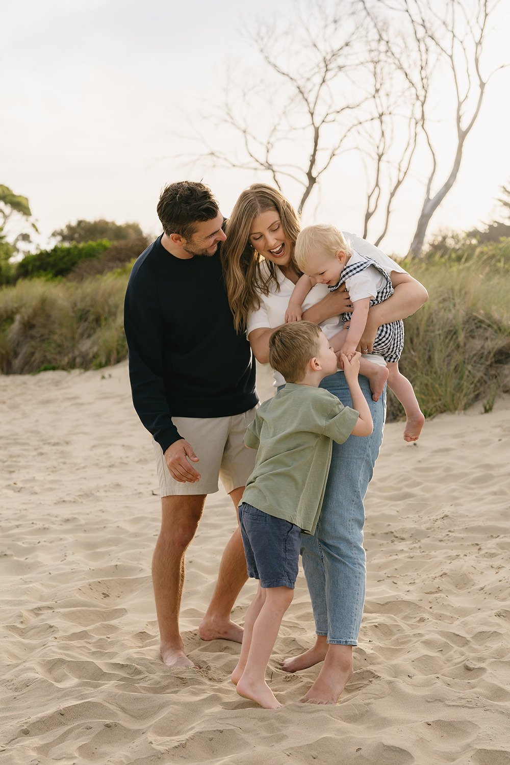 A joyful family of four playing together on a sandy beach, with trees and bushes in the background, during daylight.