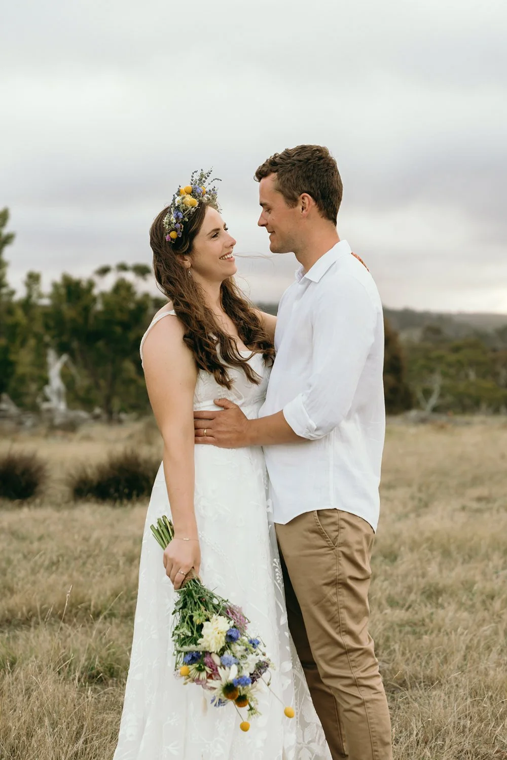 A bride and groom standing close together in an outdoor field, looking at each other and smiling during their wedding. The bride is wearing a white wedding dress and a floral crown, holding a bouquet of colorful flowers. The groom is dressed in a whi