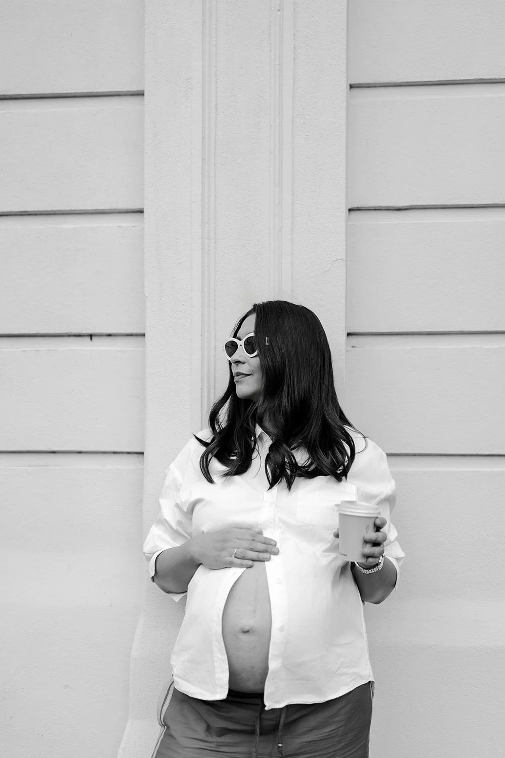 A pregnant woman with long dark hair wearing sunglasses, an open white shirt, and shorts, standing against a light-colored garage door, holding a coffee cup.