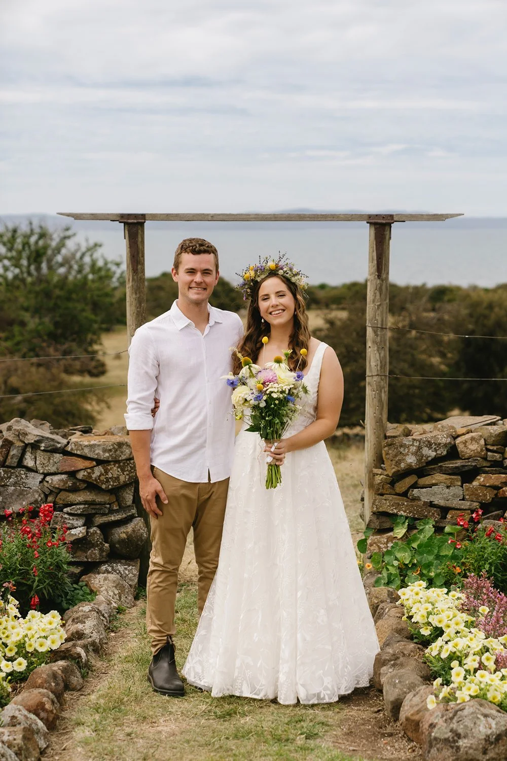A smiling couple in wedding attire standing outdoors with a stone pathway, flowers, shrubs, and a wooden pergola in the background under a cloudy sky.
