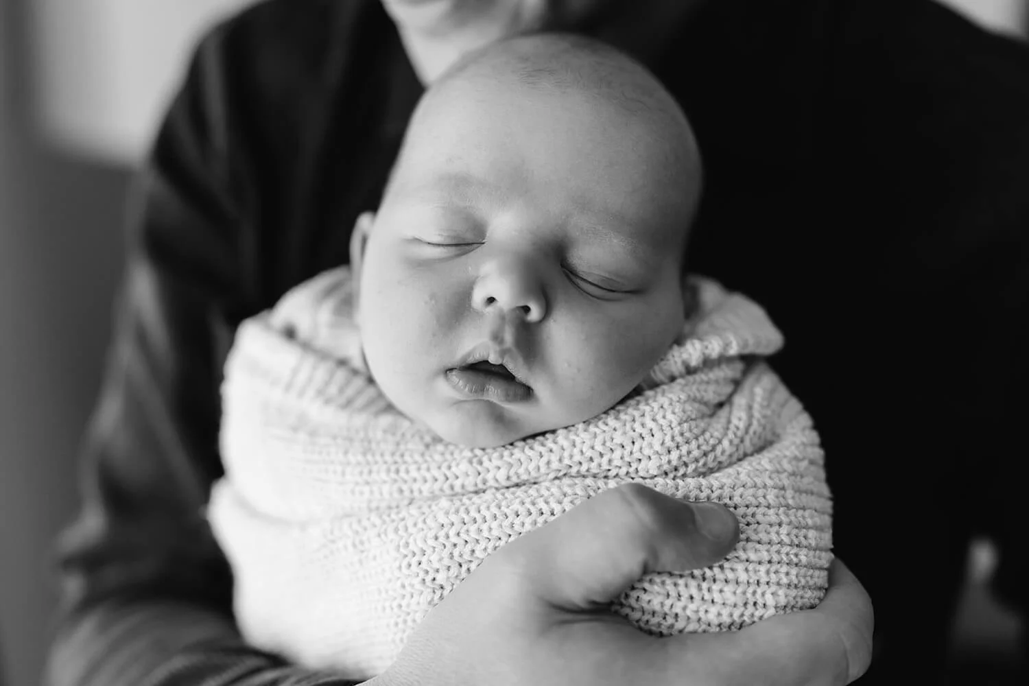 Black and white photo of a sleeping baby wrapped in a knit blanket, held gently by an adult's hand and arm.