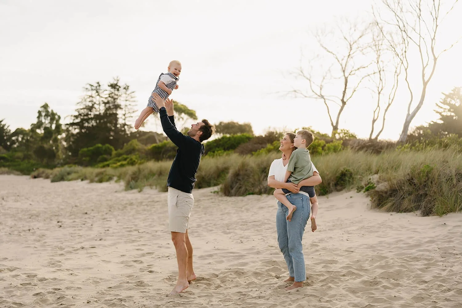 A family of four enjoying time at the beach, with the father lifting a baby in the air while the mother stands nearby holding a young boy in her arms.