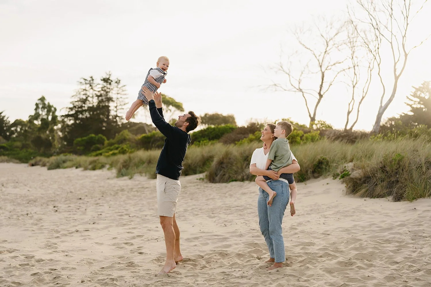 A family of four playing on a sandy beach, with a man lifting a small child into the air while a woman holds another child nearby, and trees in the background.